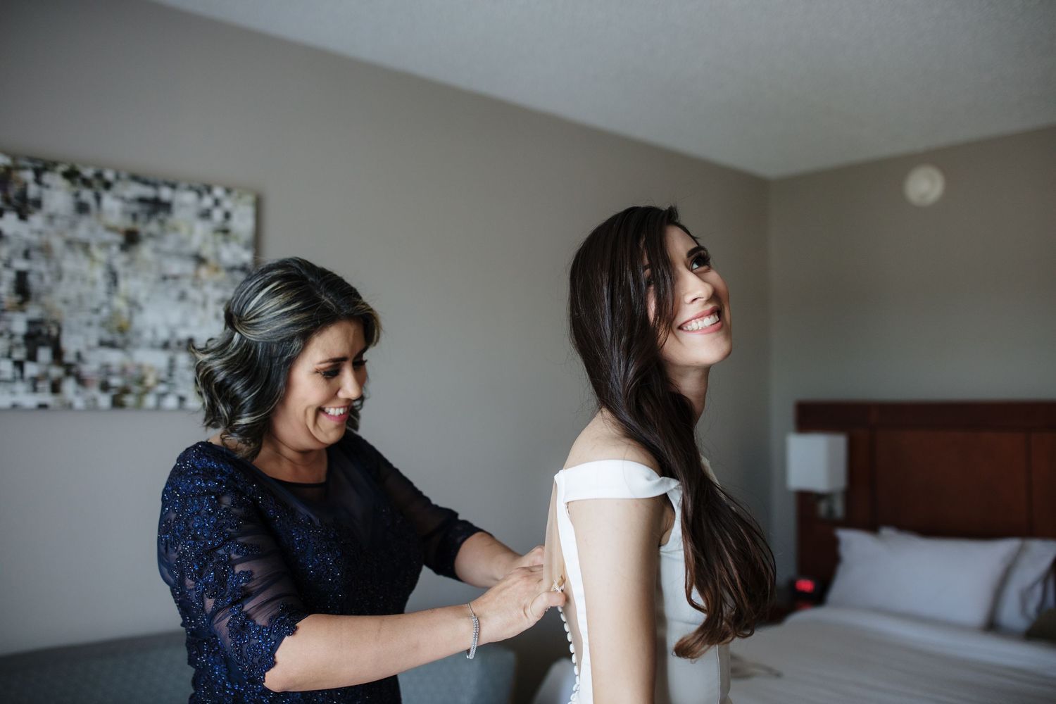 A woman helps a smiling bride button her white wedding dress in a hotel room.