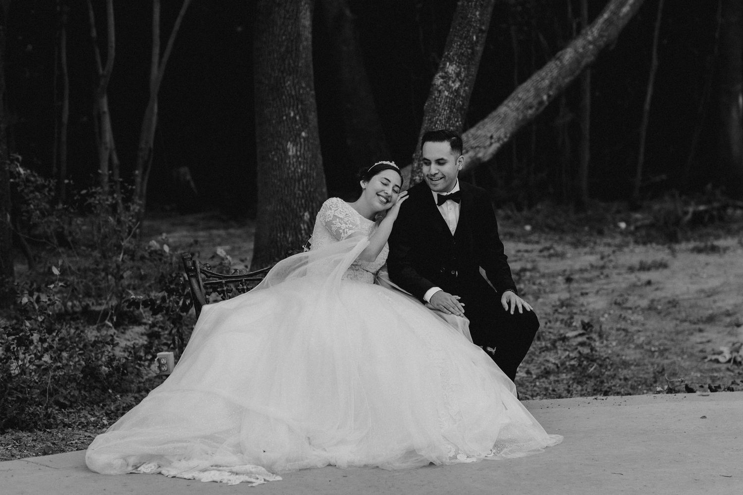 Black and white photo of bride in ball gown and groom in tuxedo sitting together outdoors among trees.