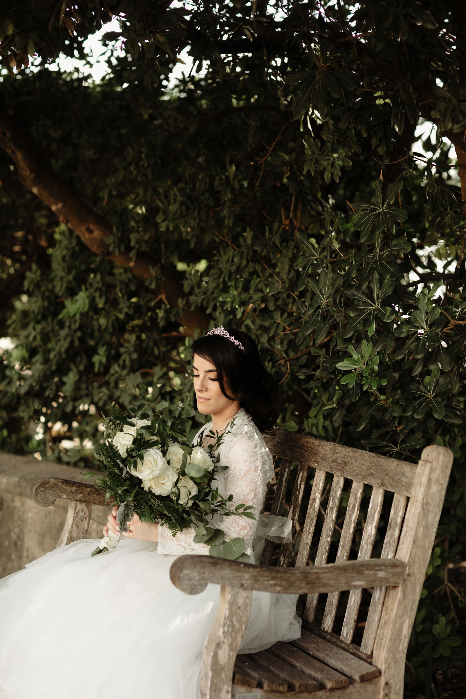 Bride in lace wedding gown sits on wooden bench holding white floral bouquet surrounded by lush green foliage.