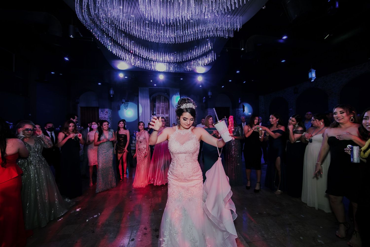 Bride in pink gown dancing joyfully with guests in elegant chandelier-lit reception hall.