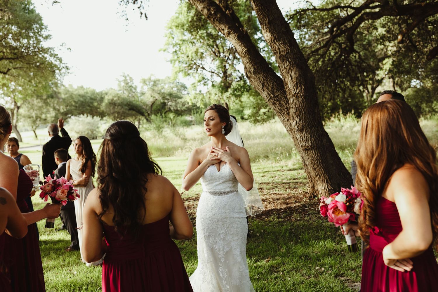 Bride in lace gown shares emotional moment with bridesmaids in red dresses outdoors.