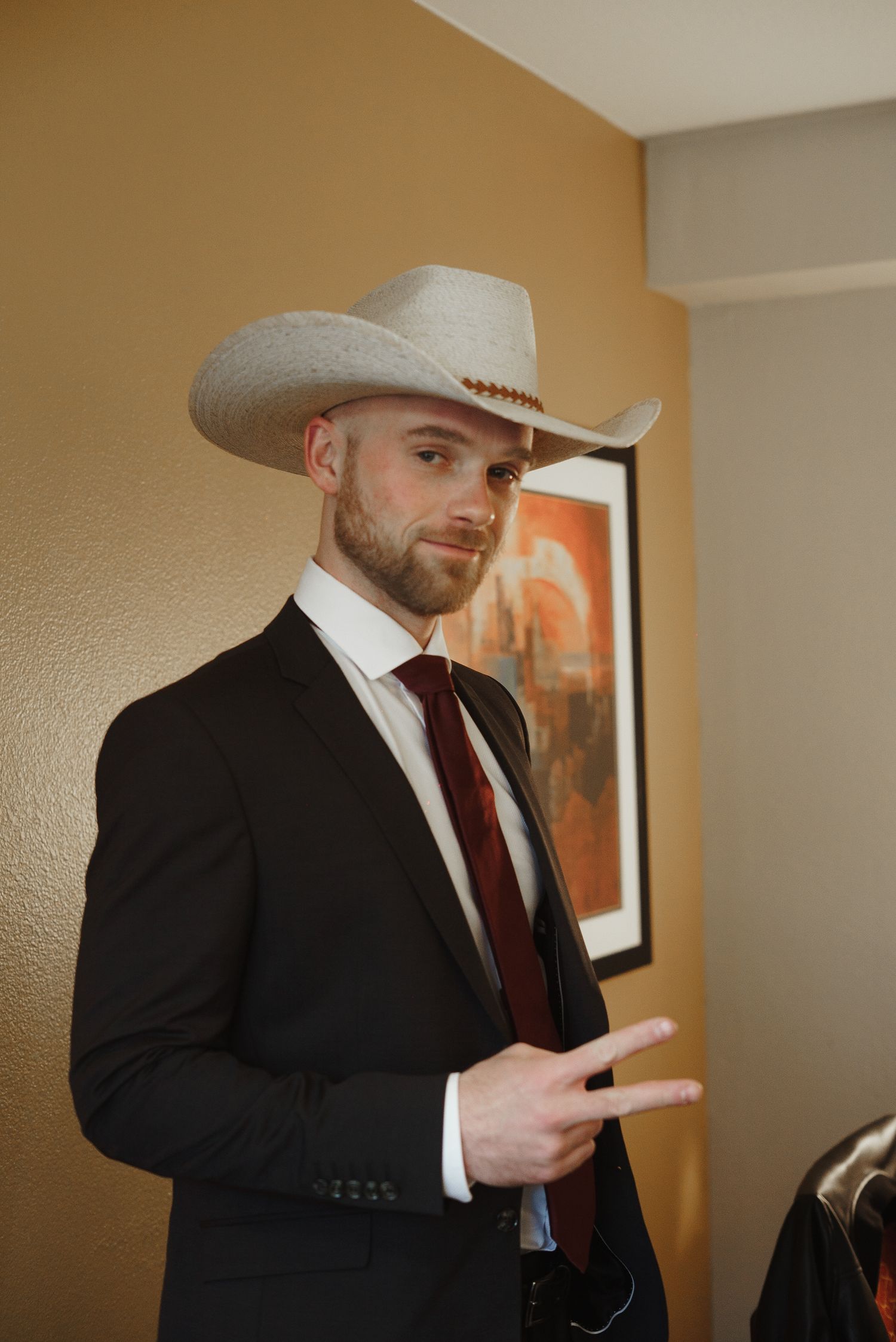 Bearded groom in black suit and white cowboy hat posing indoors before wedding.