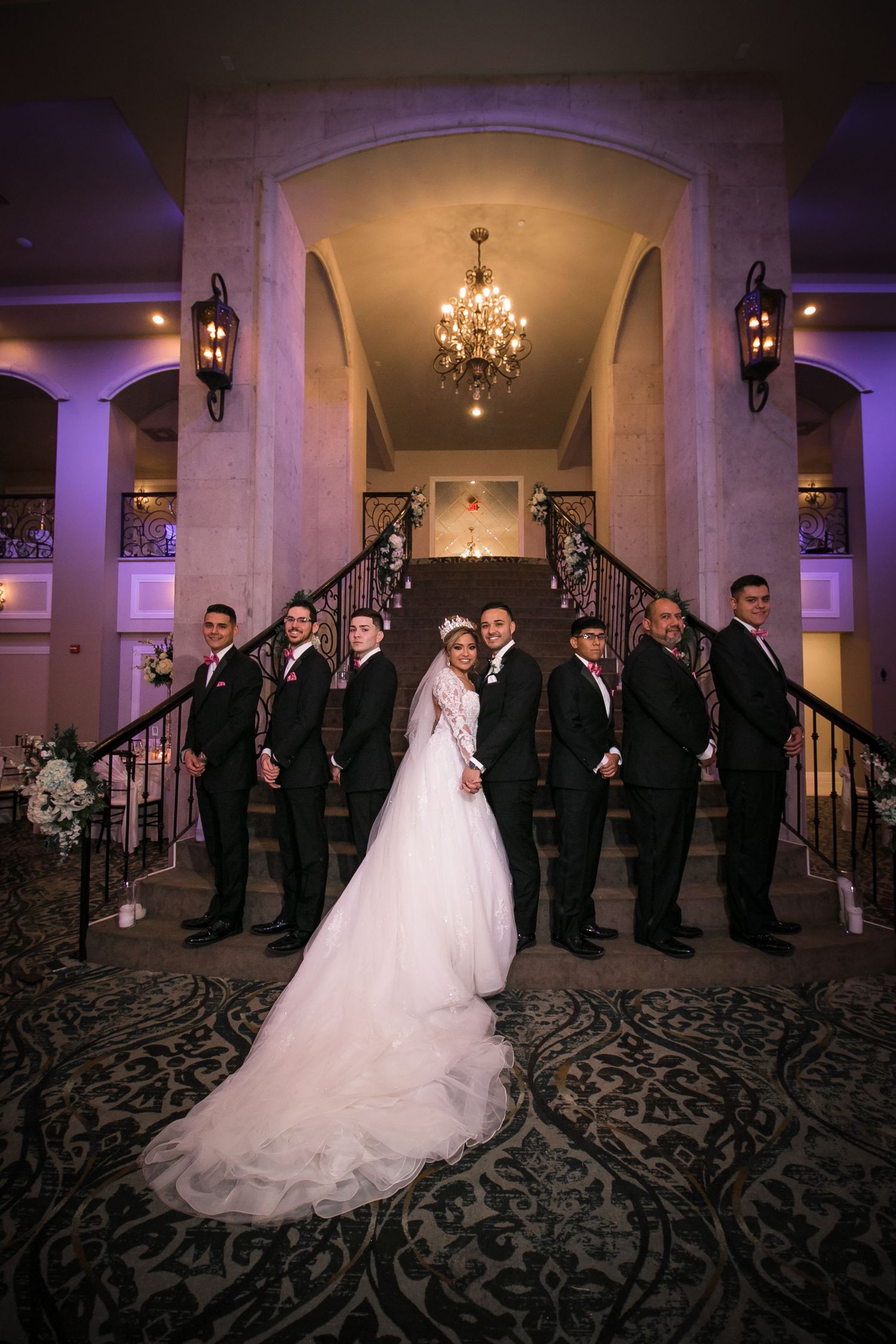 Bride in ball gown and groom with groomsmen pose elegantly on grand staircase under chandelier at reception.