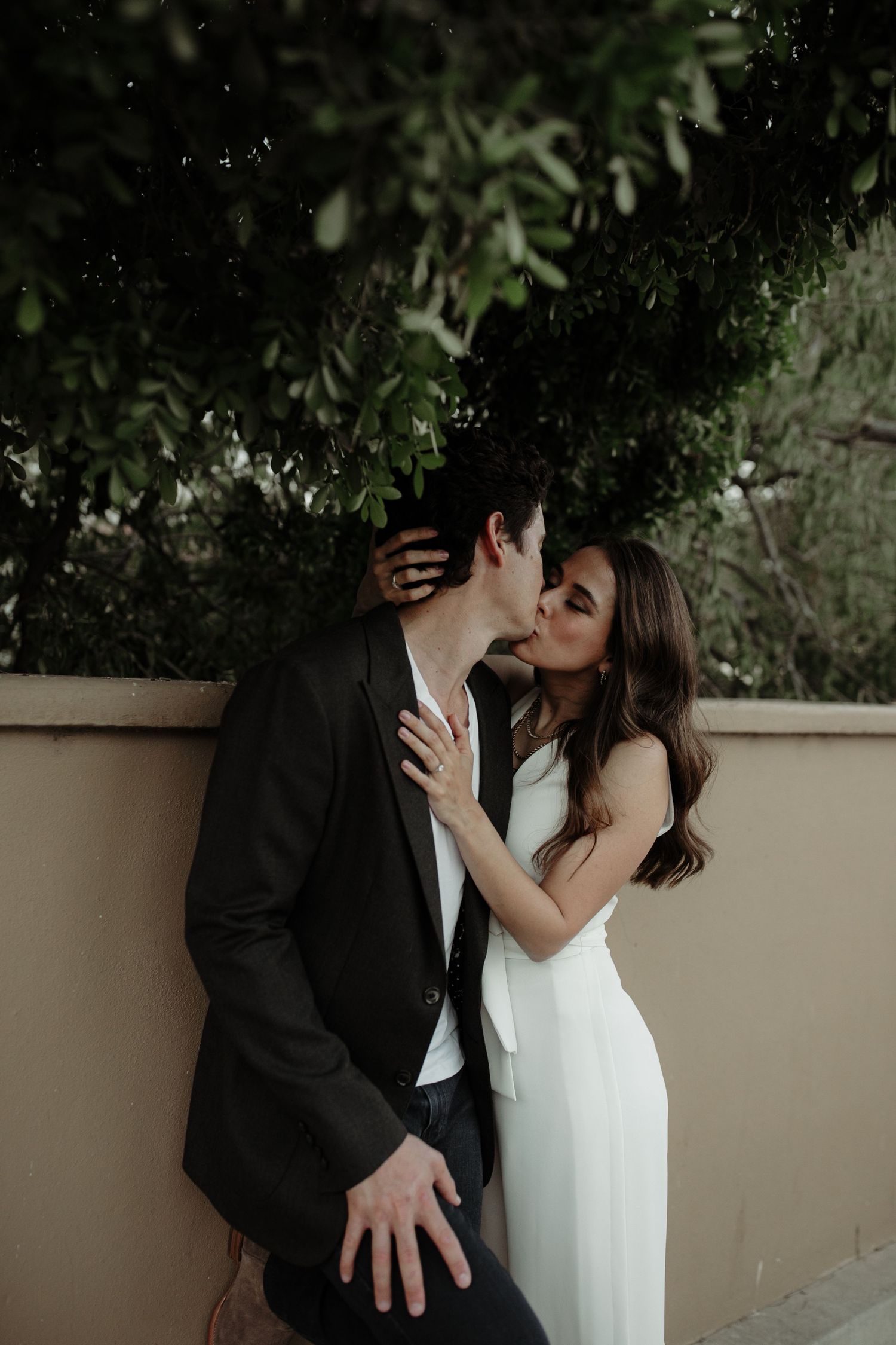 A couple shares a romantic kiss against a wall with lush greenery, dressed in elegant black and white attire.