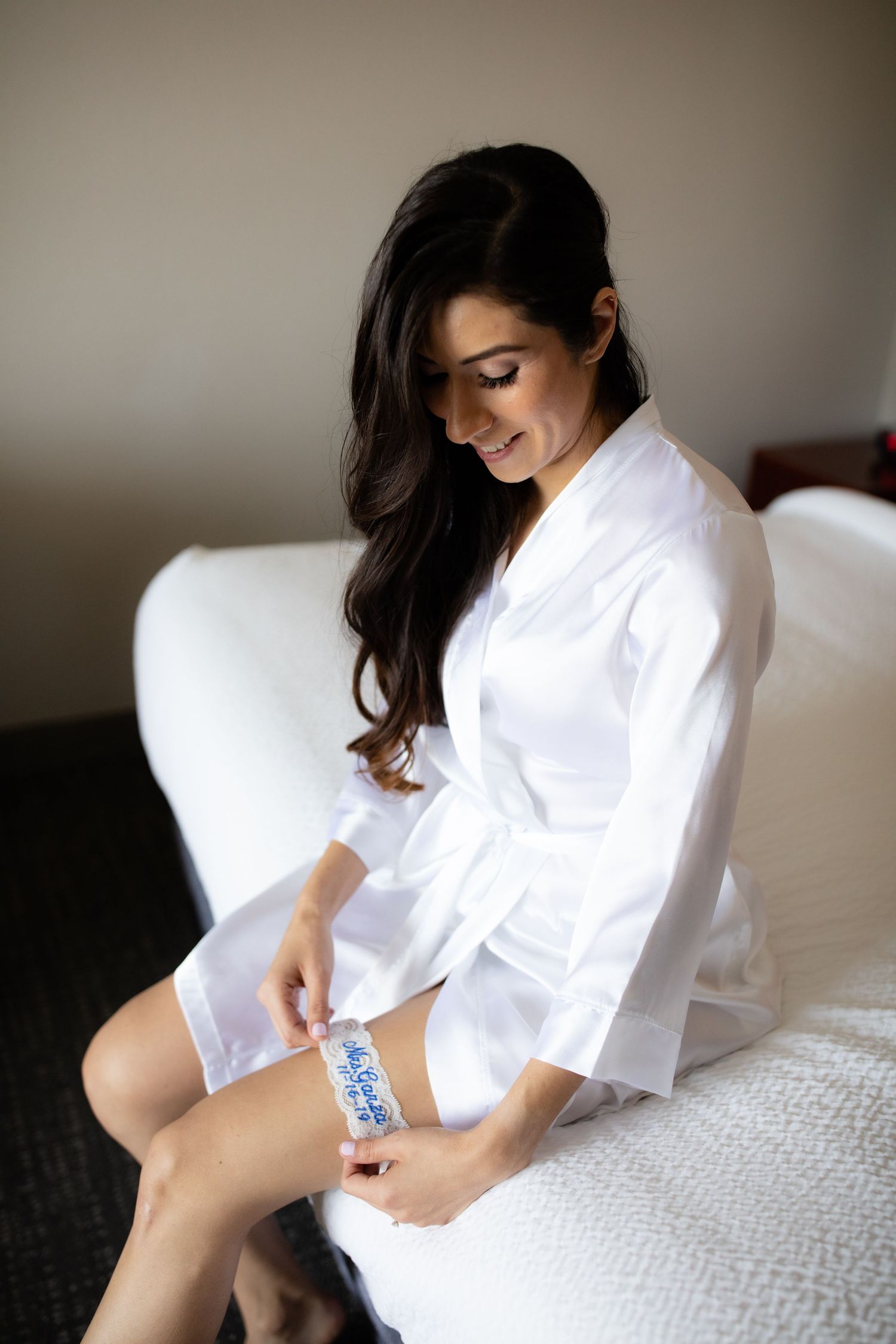 Smiling bride in white satin robe sitting on bed putting on blue wedding garter.