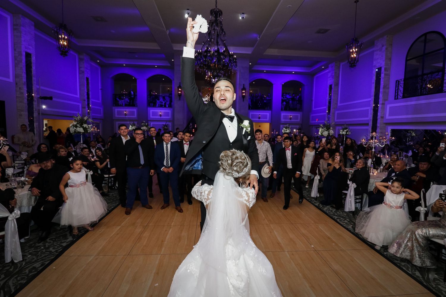 Groom lifts bride on wedding dance floor surrounded by guests in elegant ballroom with purple lighting.