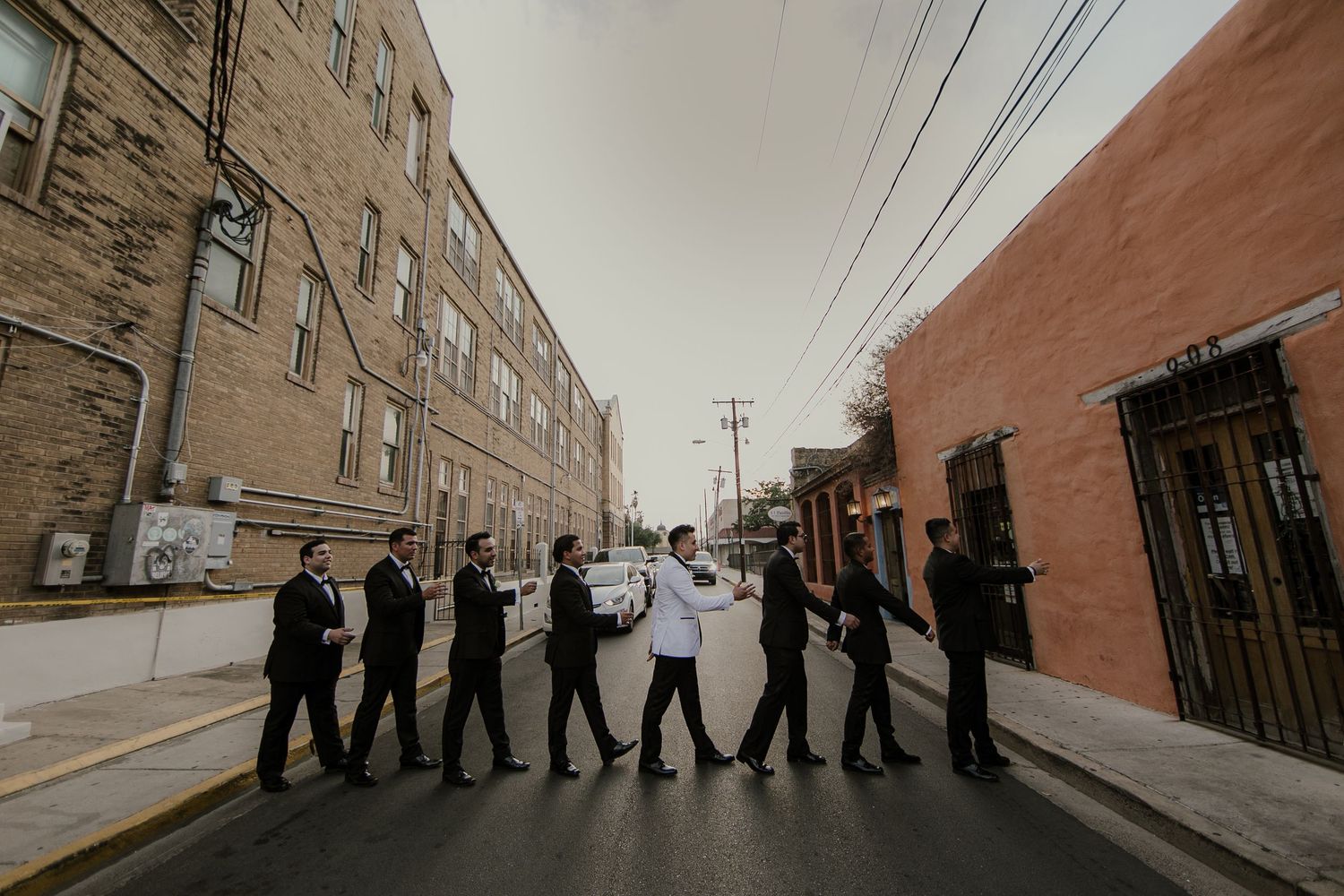 Groomsmen in black suits walk together down an urban street in a classic Abbey Road-inspired formation.