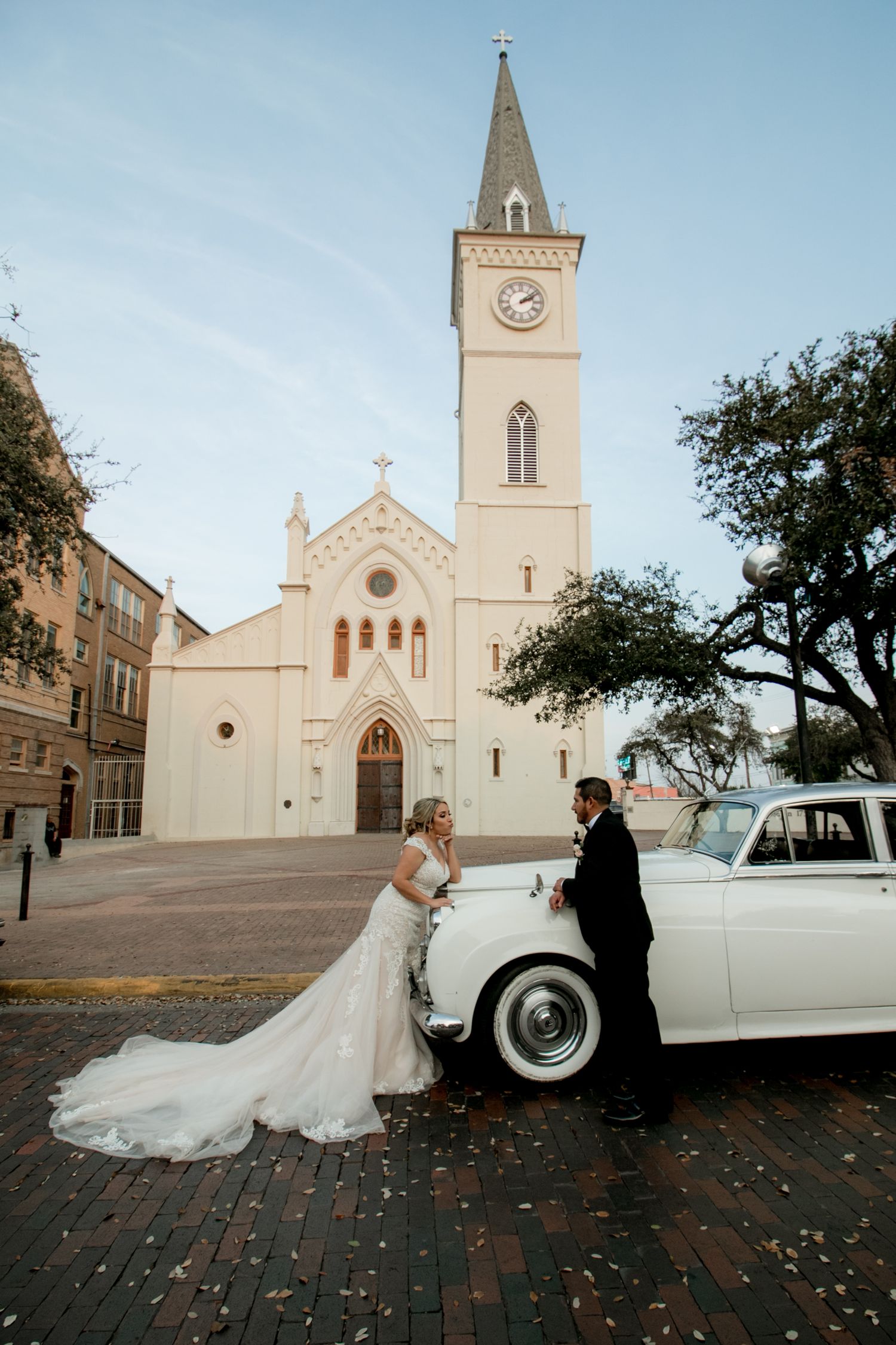 Bride and groom pose beside vintage white Rolls-Royce in front of historic church at golden hour.