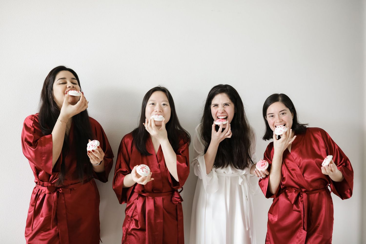 Four bridesmaids in red satin robes playfully eating macarons before the wedding ceremony.