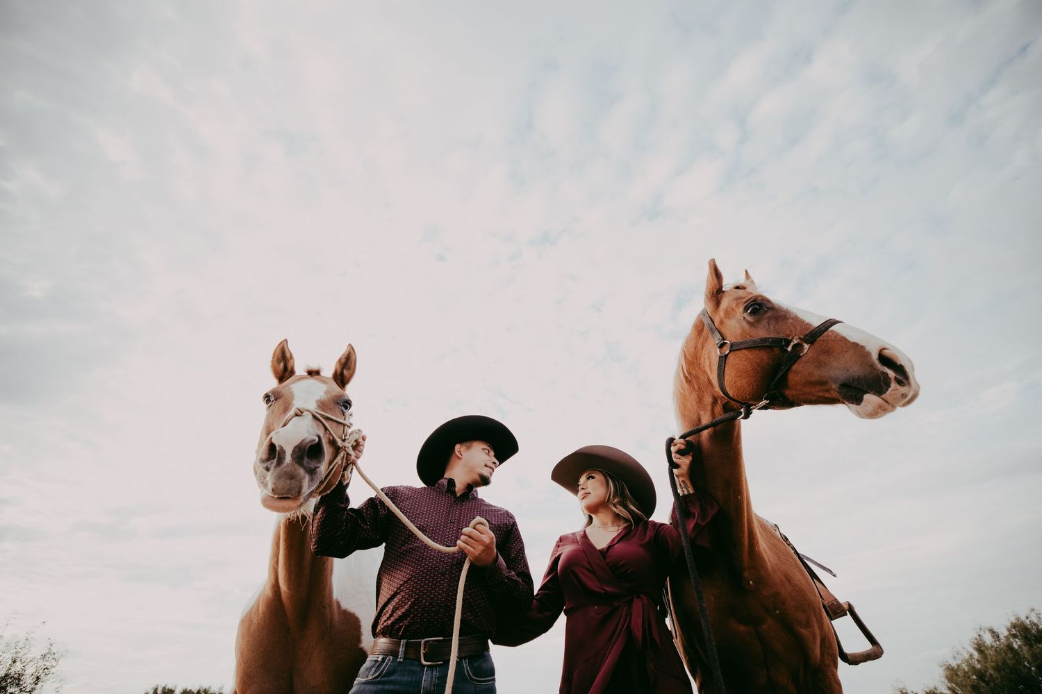Two cowboys in matching burgundy outfits and black hats hold chestnut horses under a cloudy sky.