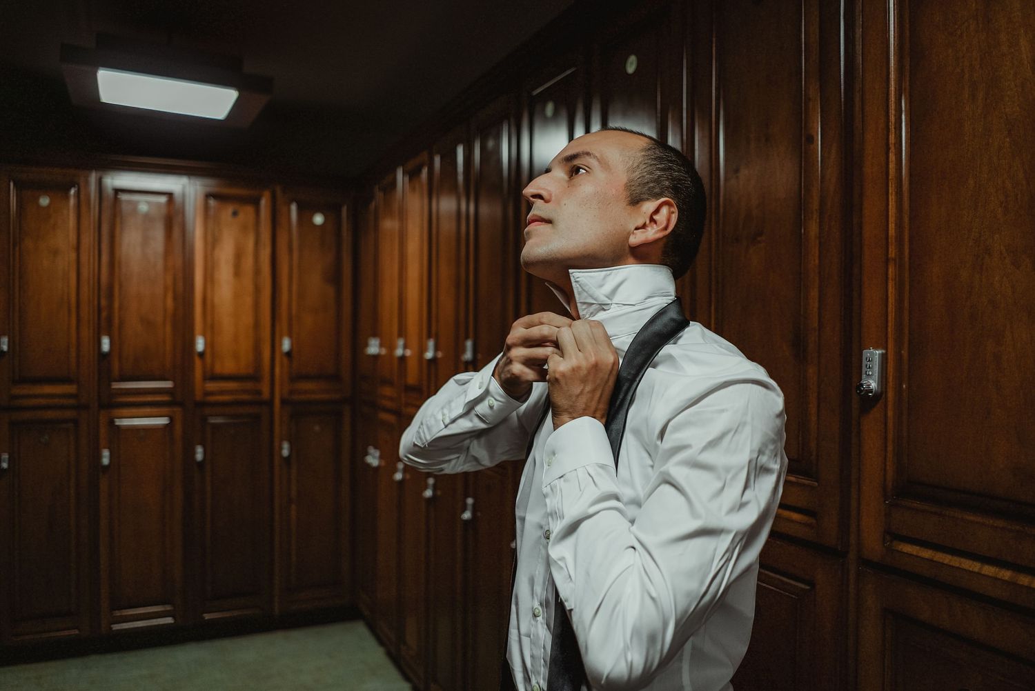 Groom in white shirt and suspenders adjusting his collar in a wooden locker room before the wedding.