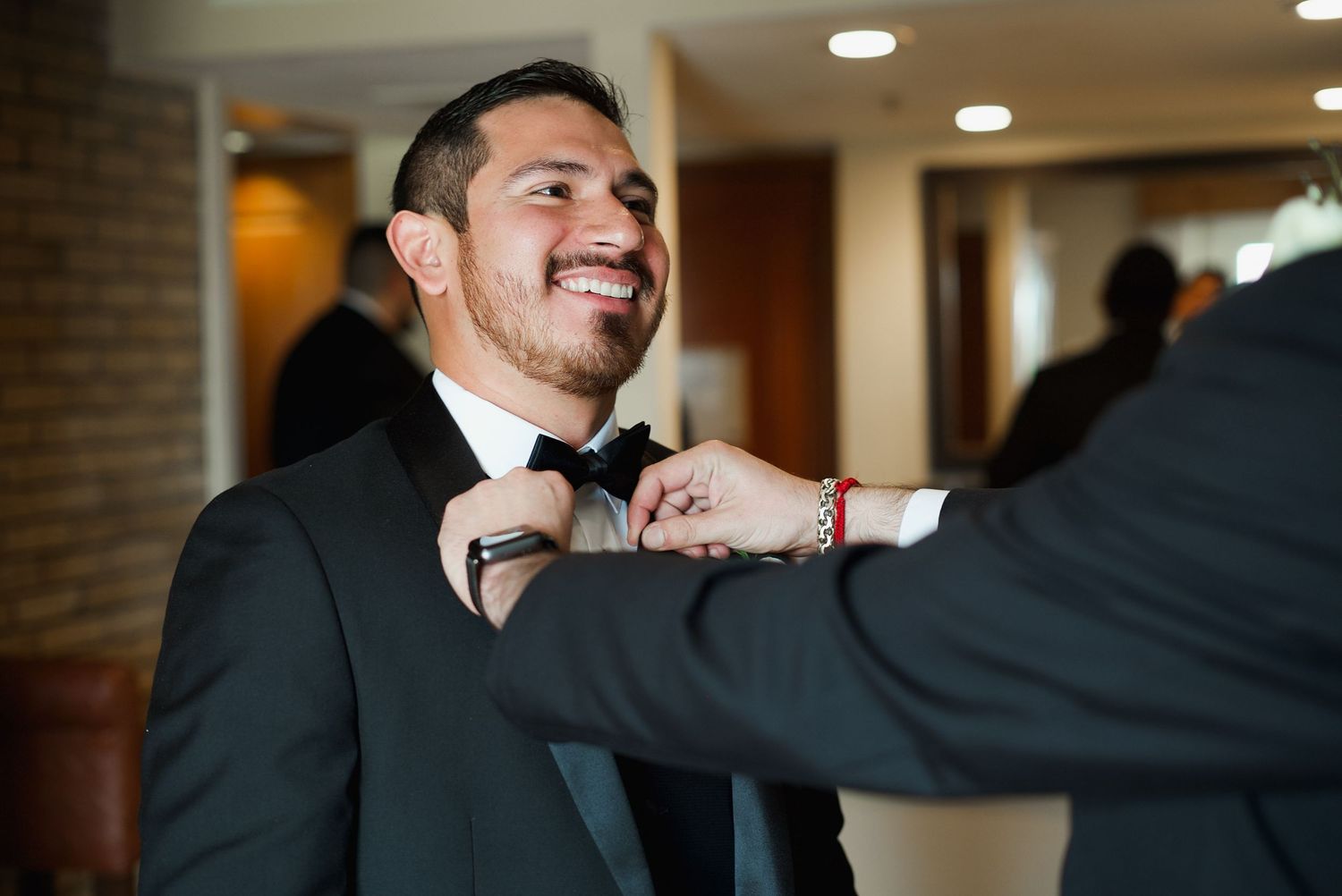 Smiling groom in black tuxedo gets bow tie adjusted by groomsman during wedding day preparations.