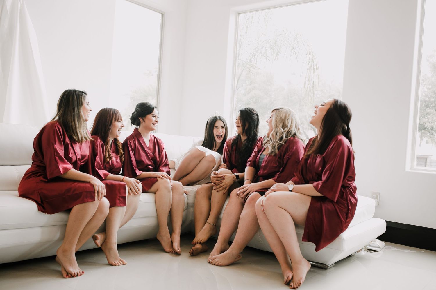 Seven bridesmaids in matching burgundy robes sitting together laughing on white sofa.