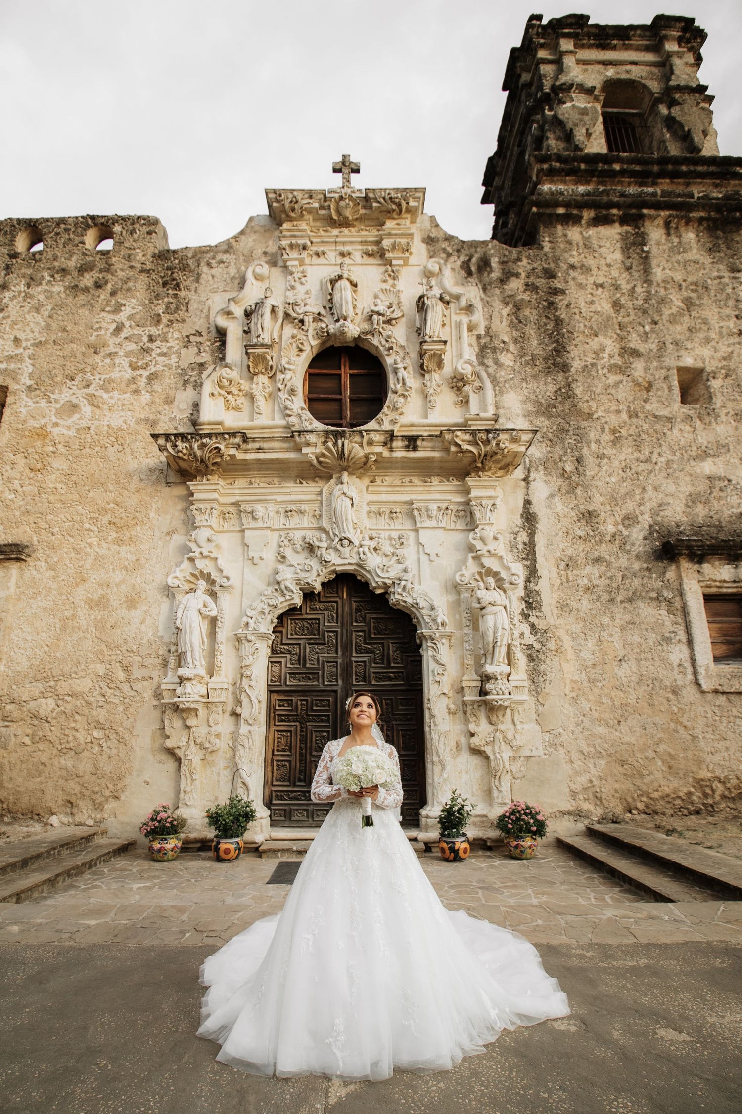 Bride in full tulle ballgown stands before elaborate baroque facade of historic San Antonio mission.