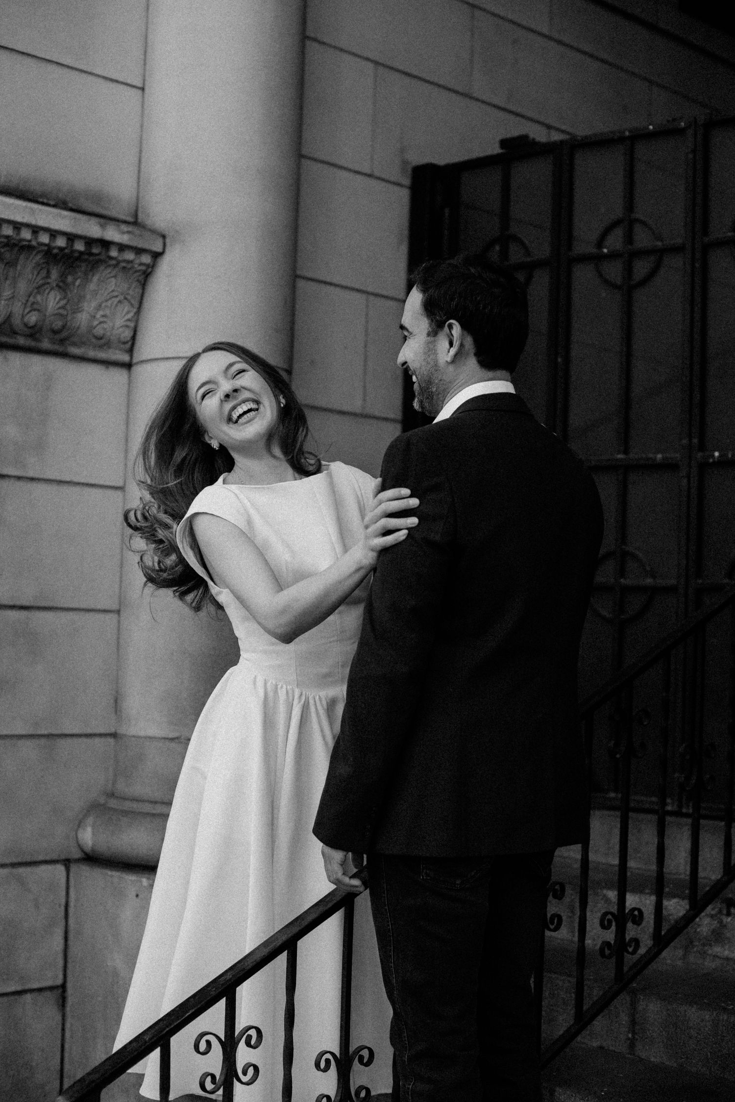 A couple shares a candid laughing moment on classic architectural steps in black and white.