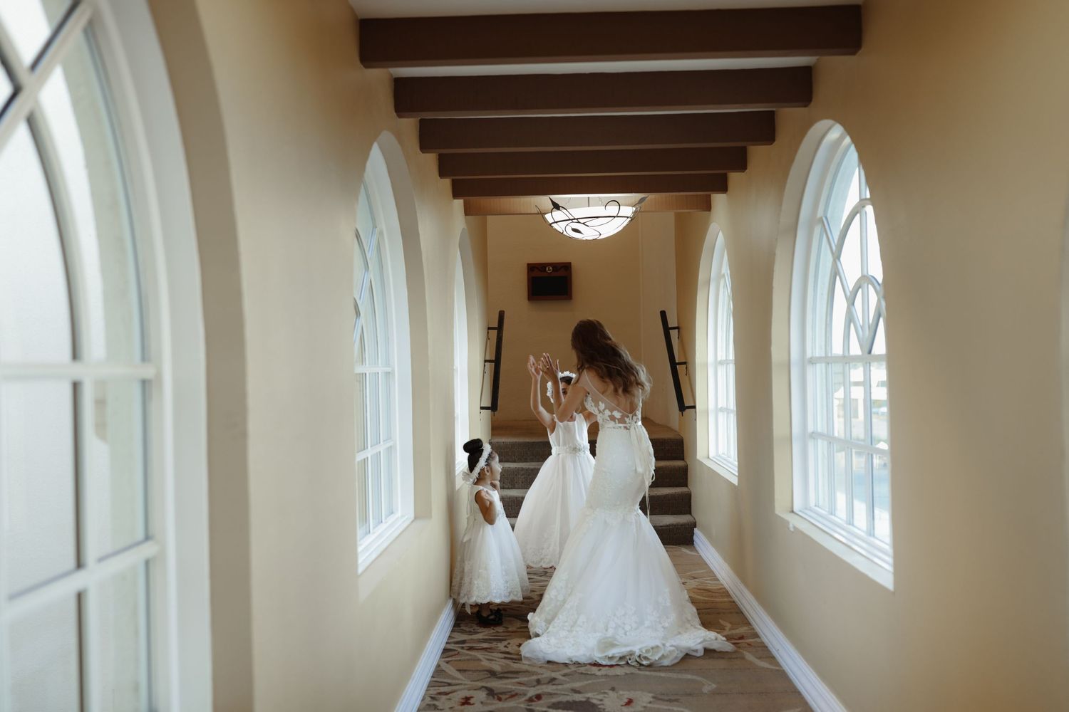 Bride in fitted gown walks with flower girl in white dress down elegant hallway with arched windows.