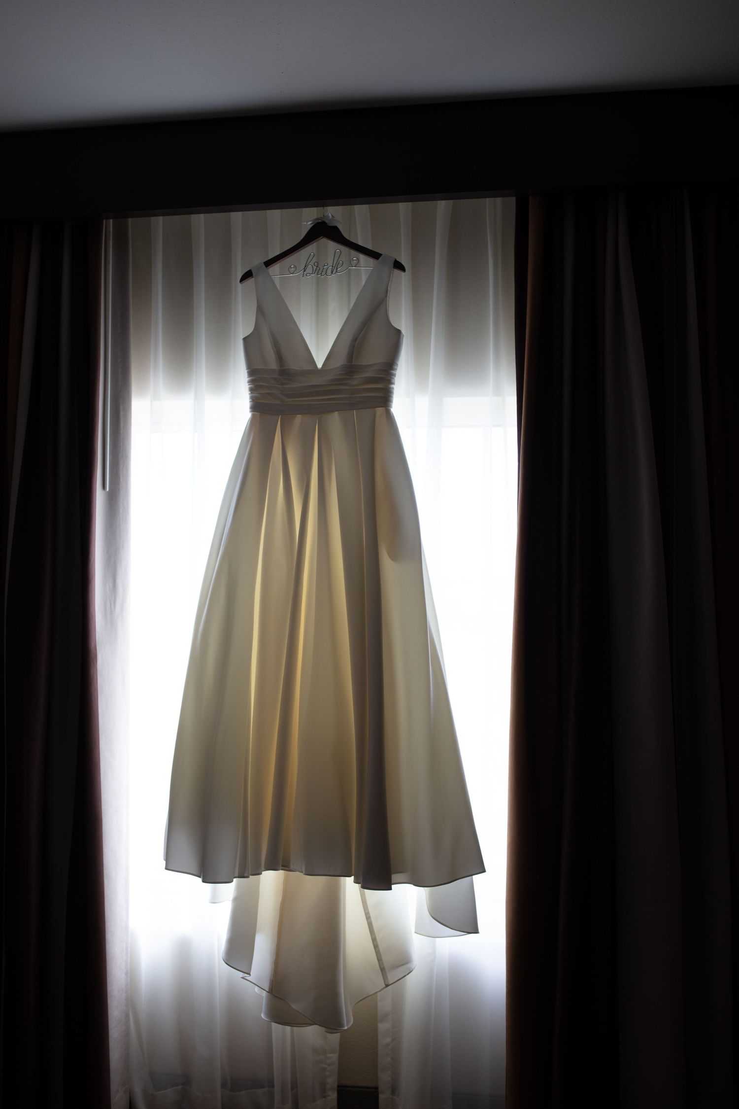Elegant champagne-colored wedding gown hanging on a hanger in front of a bright window.