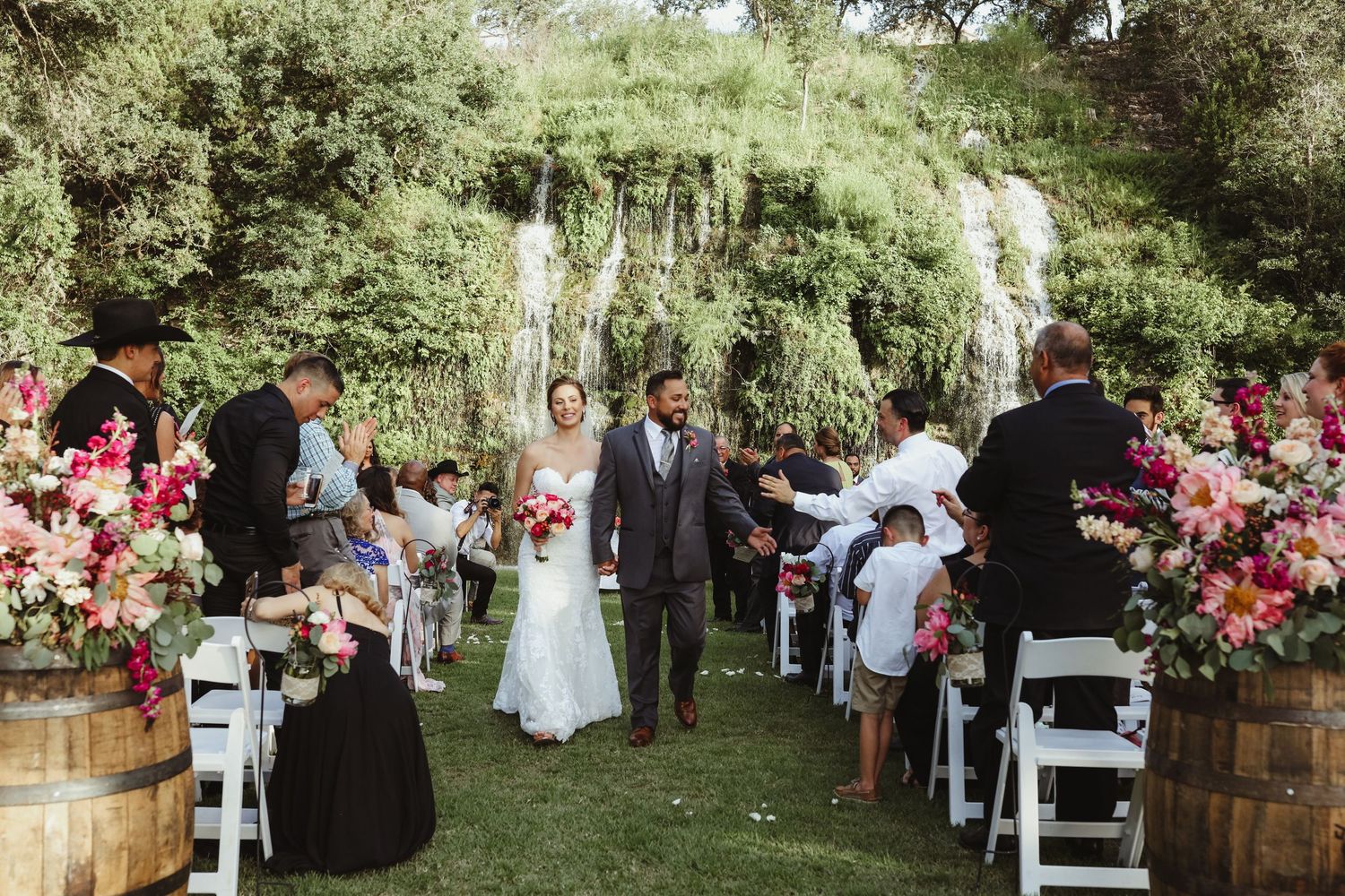 Newlyweds walk down aisle surrounded by vibrant pink floral arrangements at garden wedding.