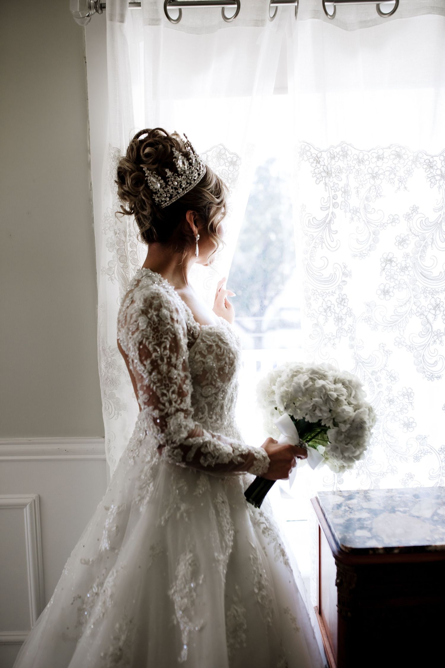 Elegant bride in lace gown and crystal crown gazes out a window holding white bouquet.