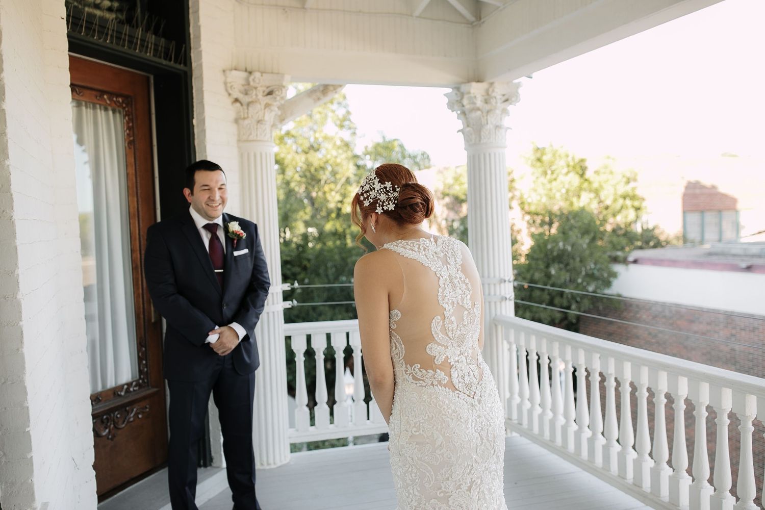 Groom in navy suit sees bride in lace backless wedding dress for first time on white columned porch.