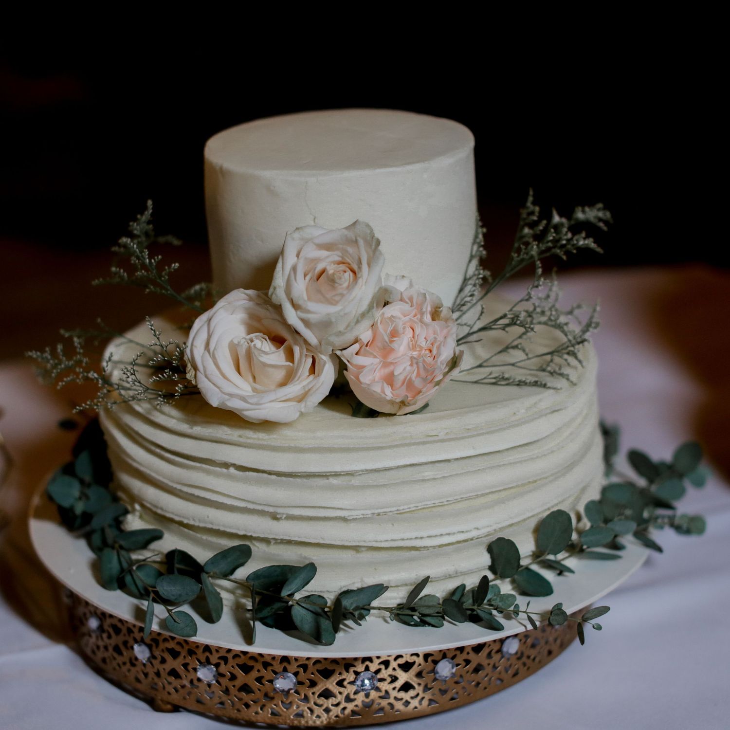 Two-tier white textured wedding cake adorned with blush roses and eucalyptus greenery on gold cake stand.