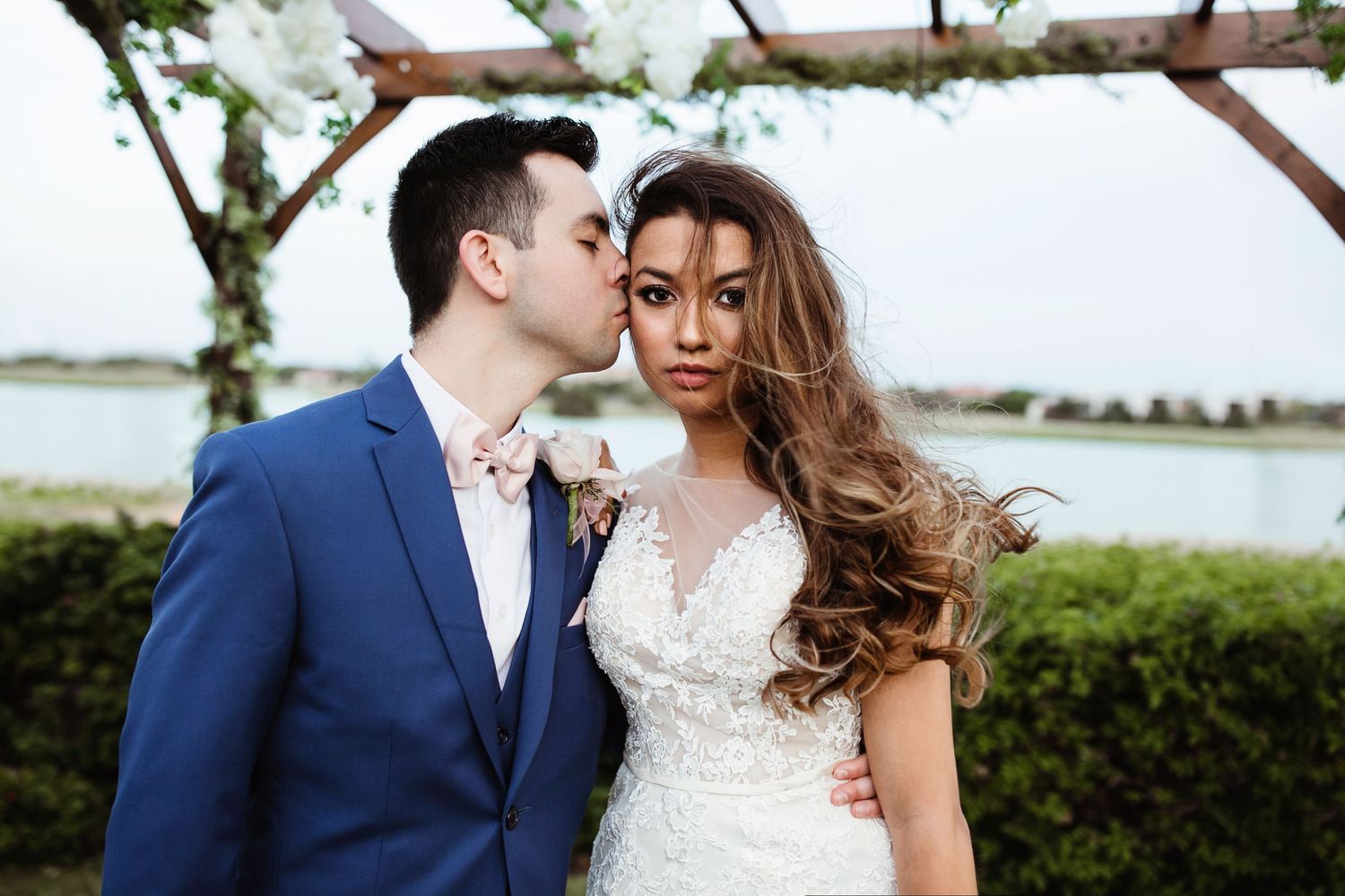 Groom in blue suit kisses bride in lace wedding dress outdoors near waterfront.