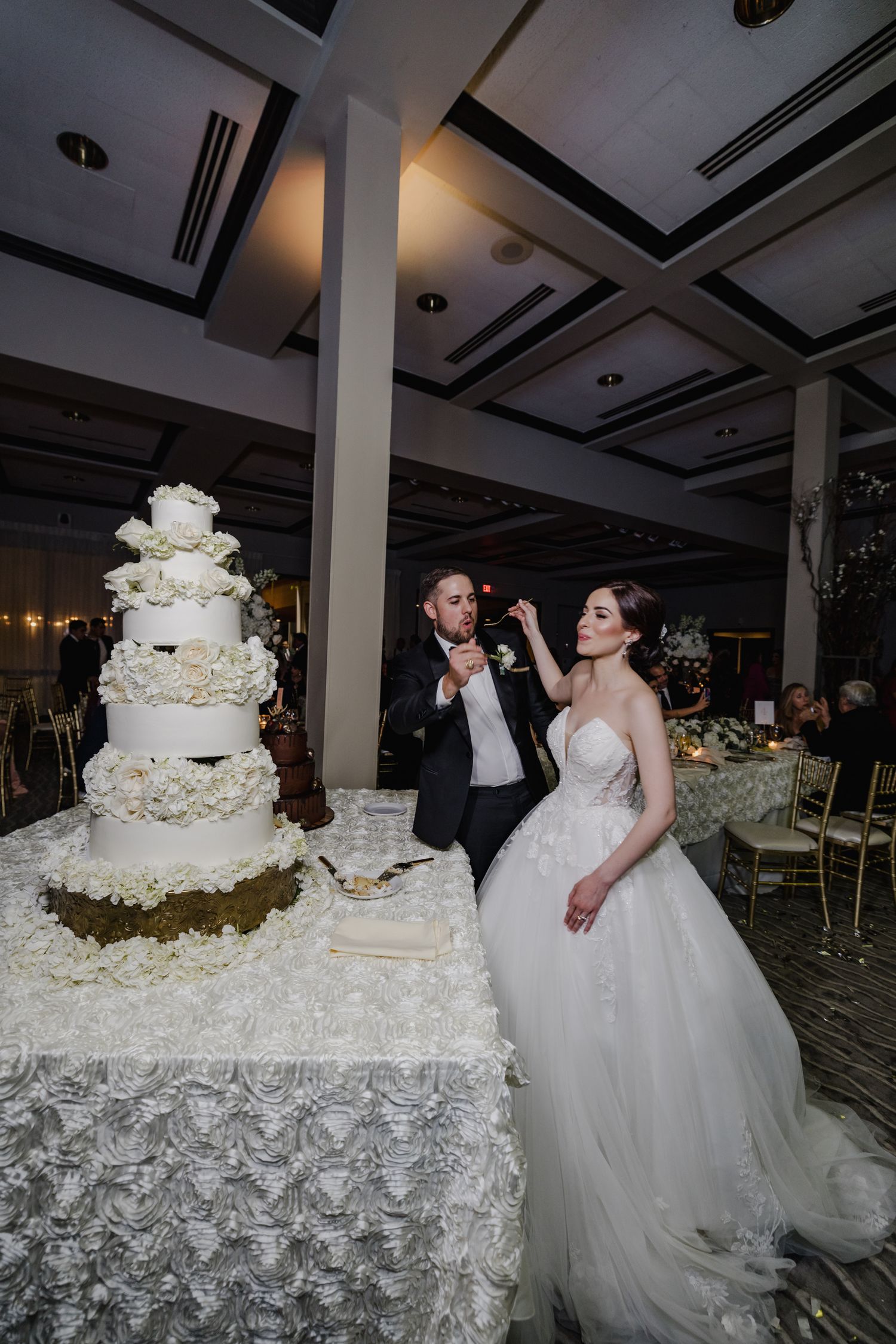 Bride and groom cutting their elegant white five-tier wedding cake adorned with floral decorations at a formal reception.