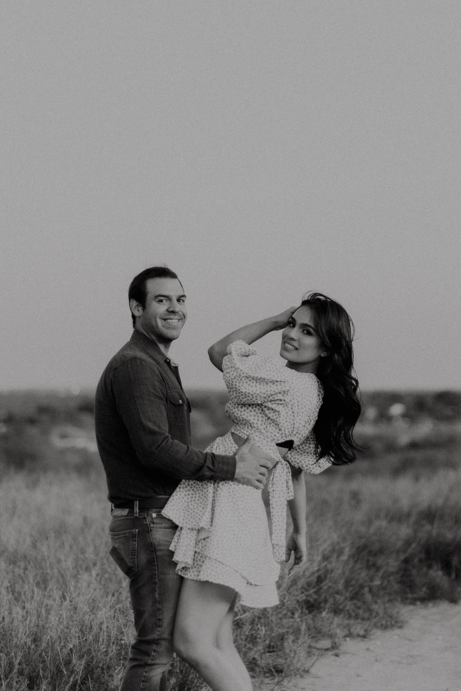 Black and white photo of smiling couple embracing in open field at dusk.