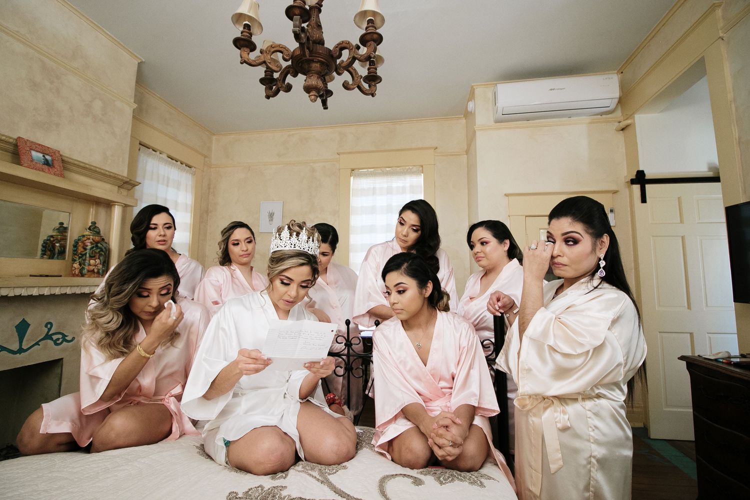 Bridesmaids in satin robes gather around bride reading heartfelt letter in vintage bridal suite.
