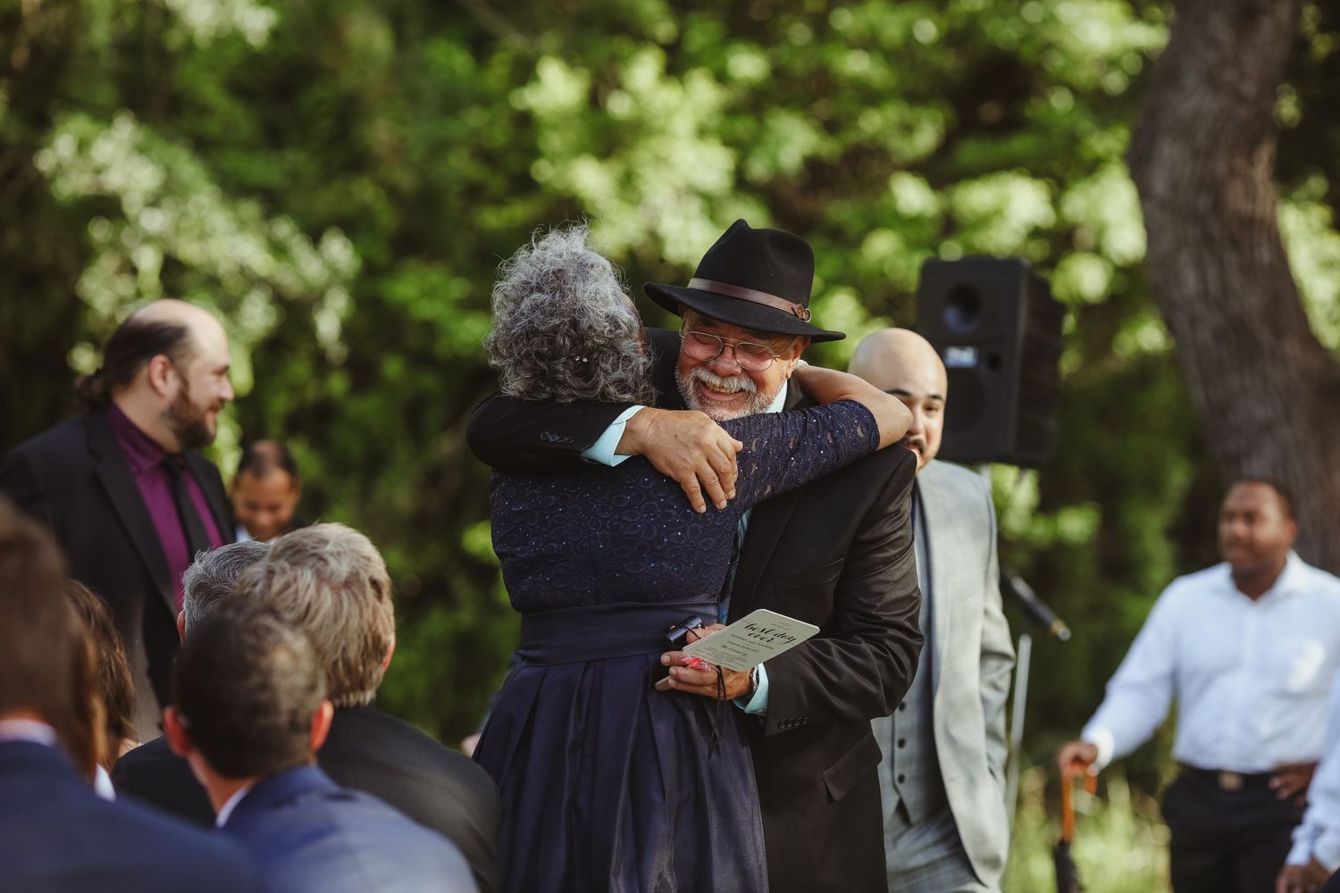 Two guests share a warm embrace during an outdoor wedding ceremony surrounded by lush green trees.
