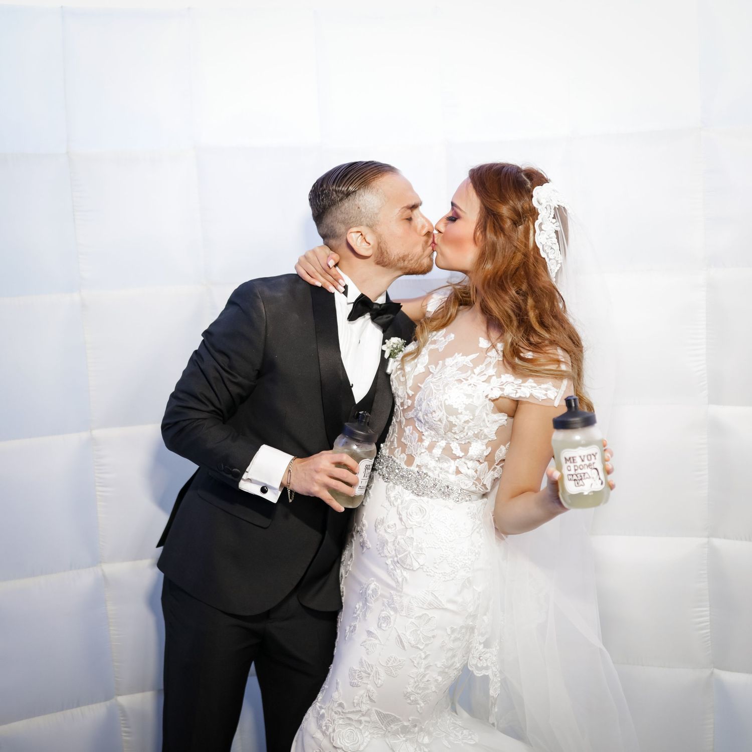 Newlywed couple sharing a kiss at a wedding photo booth holding mason jar drinks.