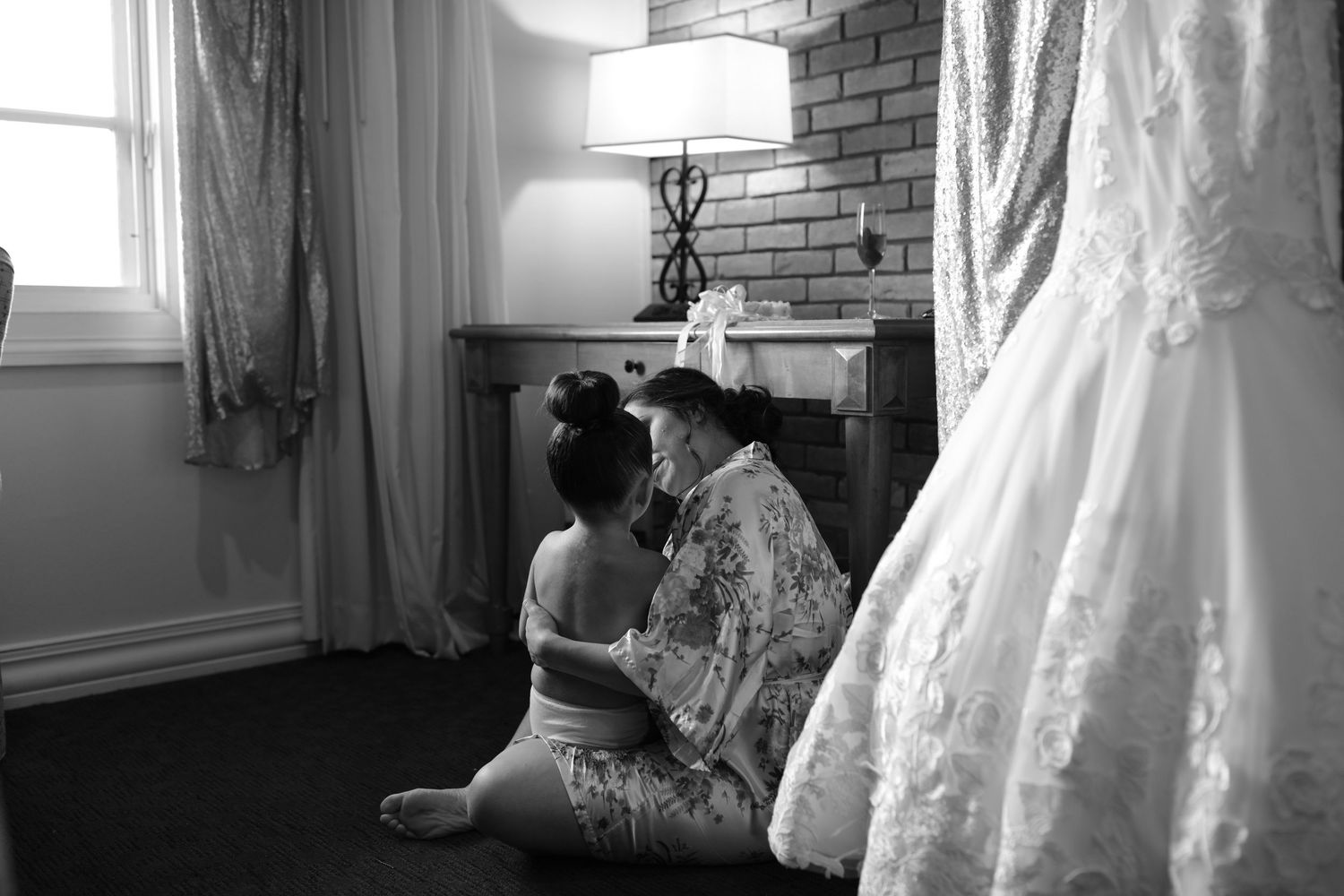 Black and white photo of bride sitting with attendant near fireplace beside hanging wedding gown.