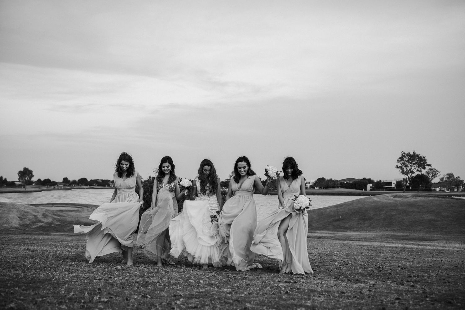 Black and white photo of bride and five bridesmaids in flowing gowns walking across open field.