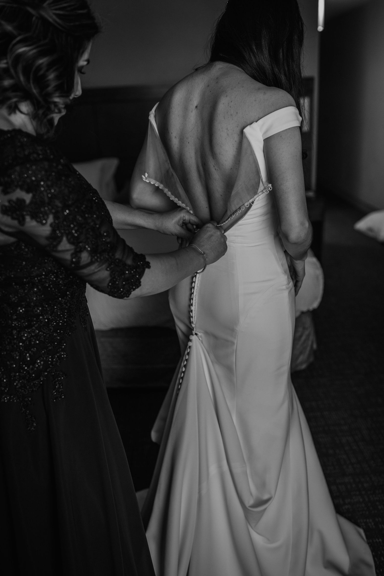 Black and white photo of a bride having her wedding dress buttoned up the back by a bridesmaid.