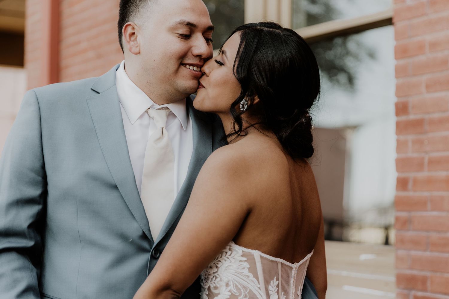 Intimate moment between bride and groom sharing a kiss against brick wall backdrop.