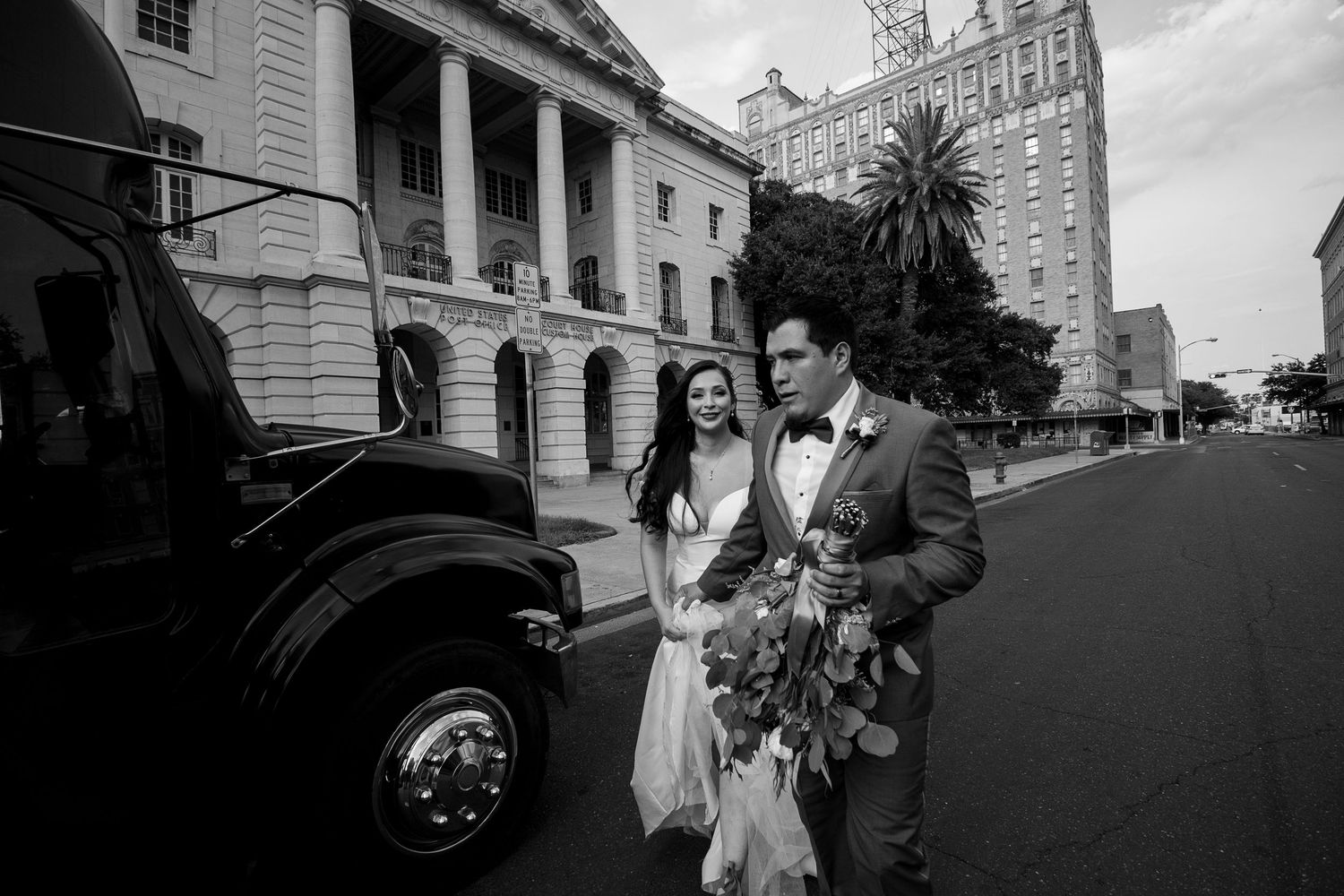 Black and white photo of bride and groom walking past vintage truck near grand building.