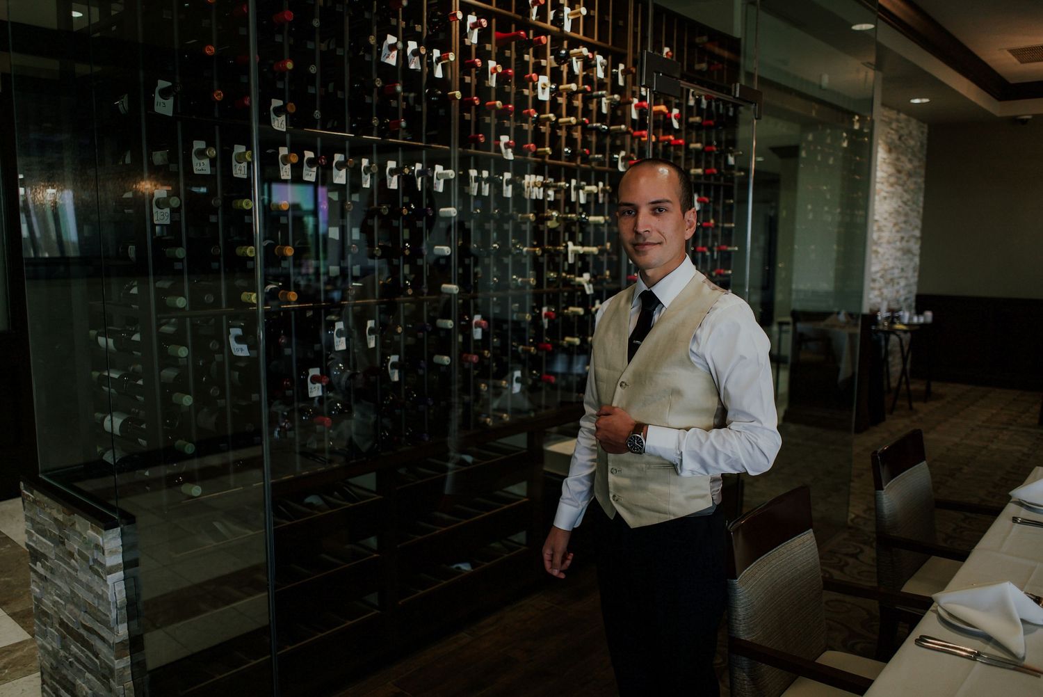 Elegant waiter in vest standing by wine wall display in upscale restaurant.