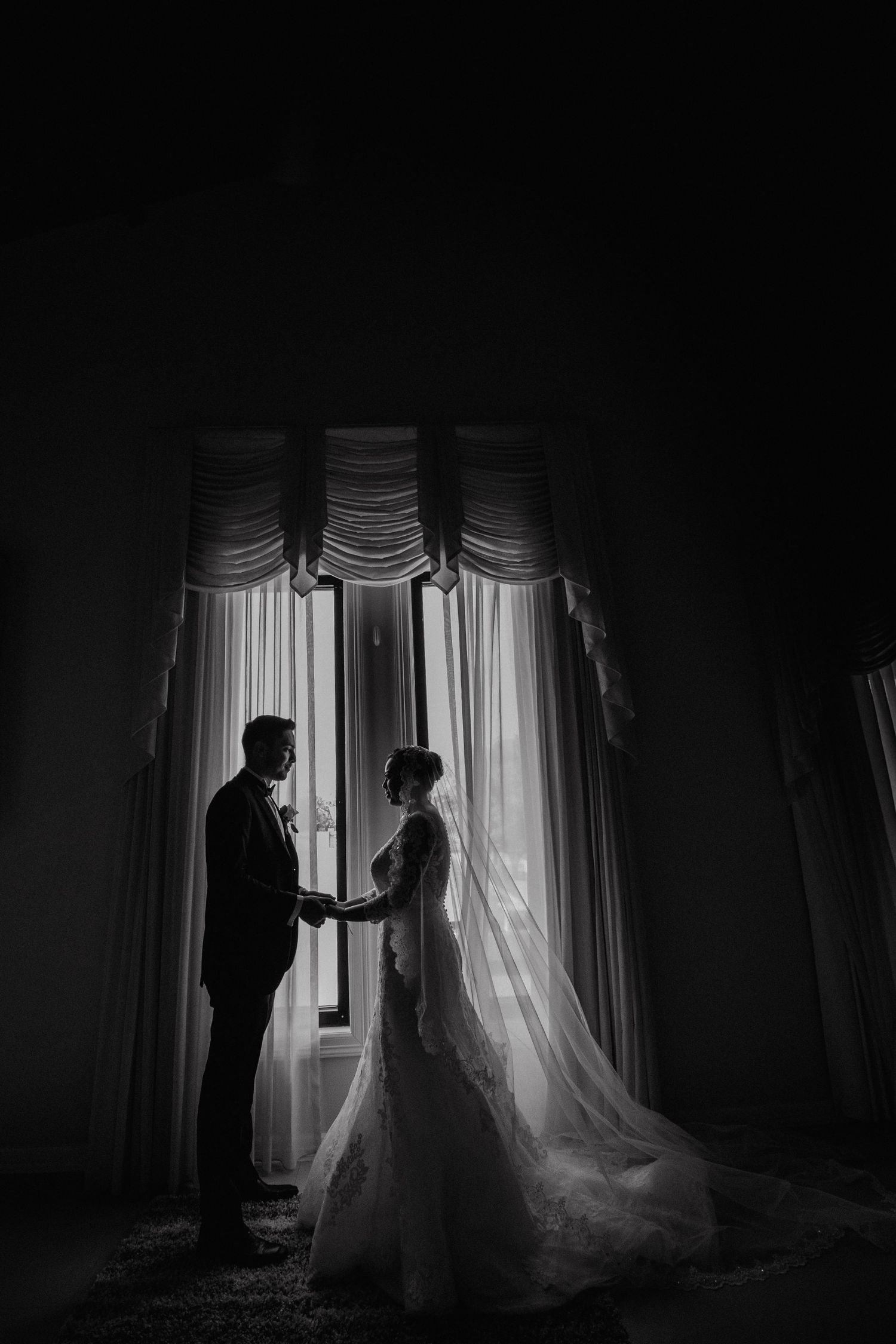 Silhouette of bride and groom holding hands by draped window in romantic black and white photo.