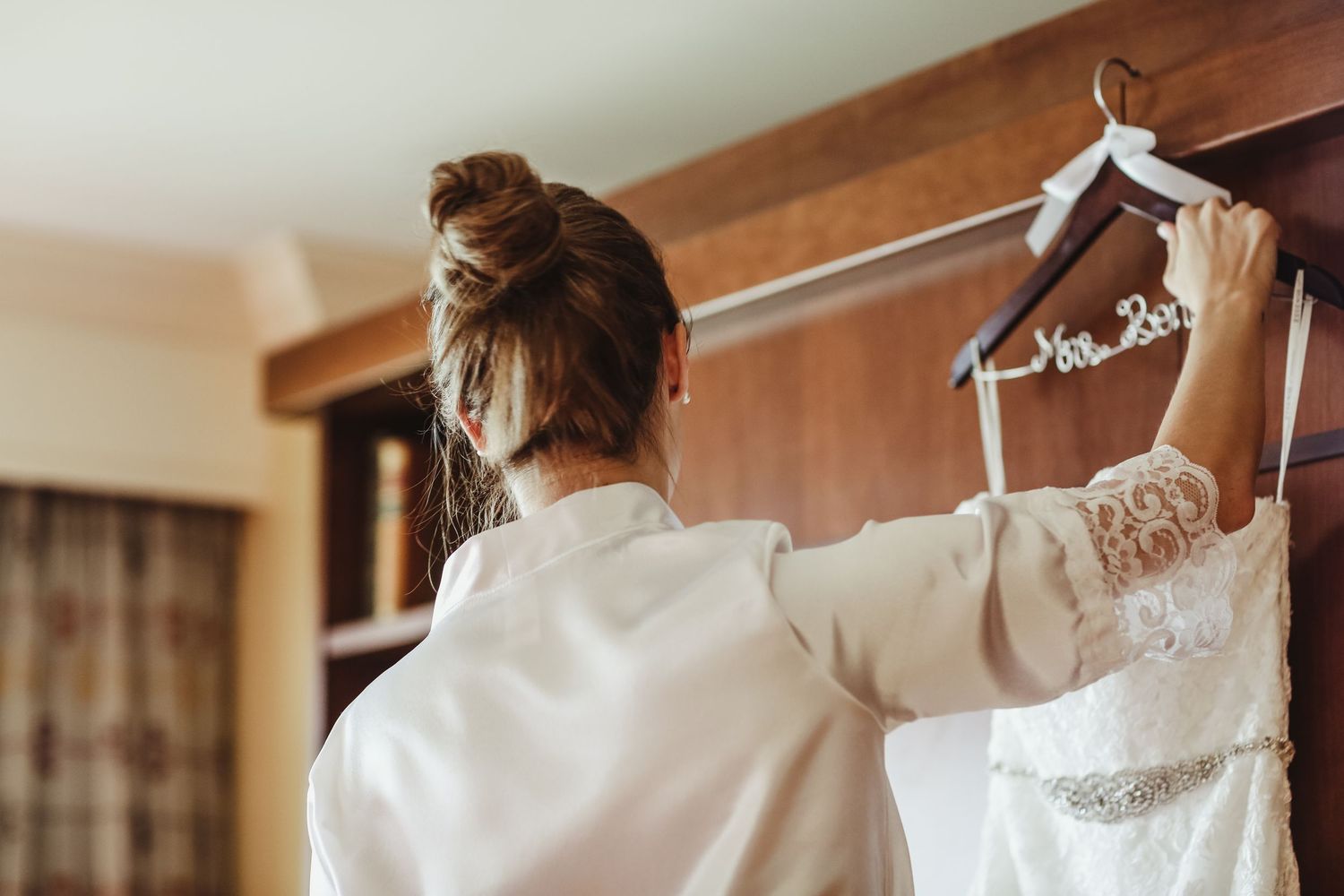 Bride in white robe reaching for wedding dress on hanger labeled Mrs. on wooden rack.