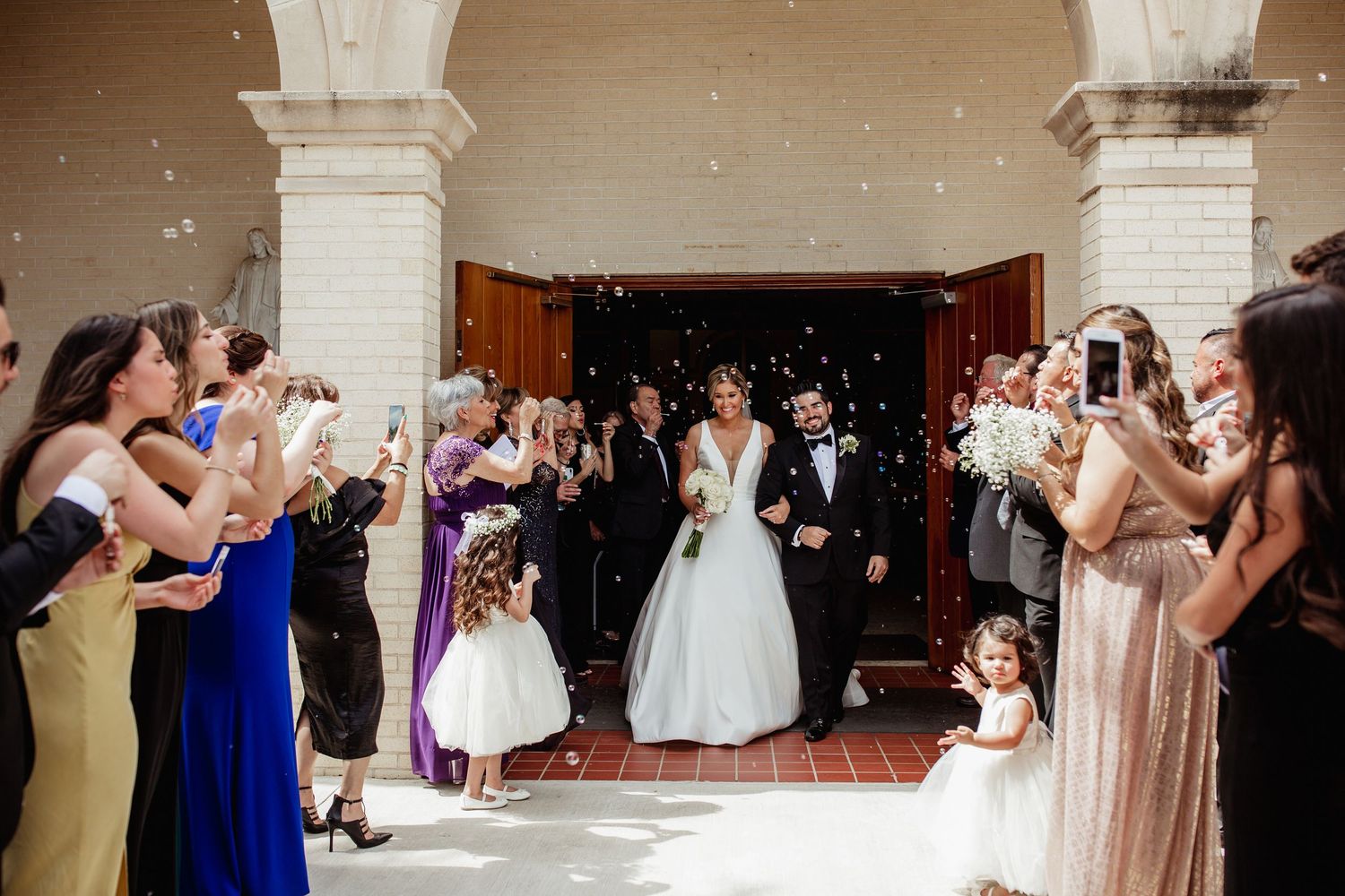 Newlyweds exit church ceremony as guests toss confetti while flower girls watch the joyful celebration.