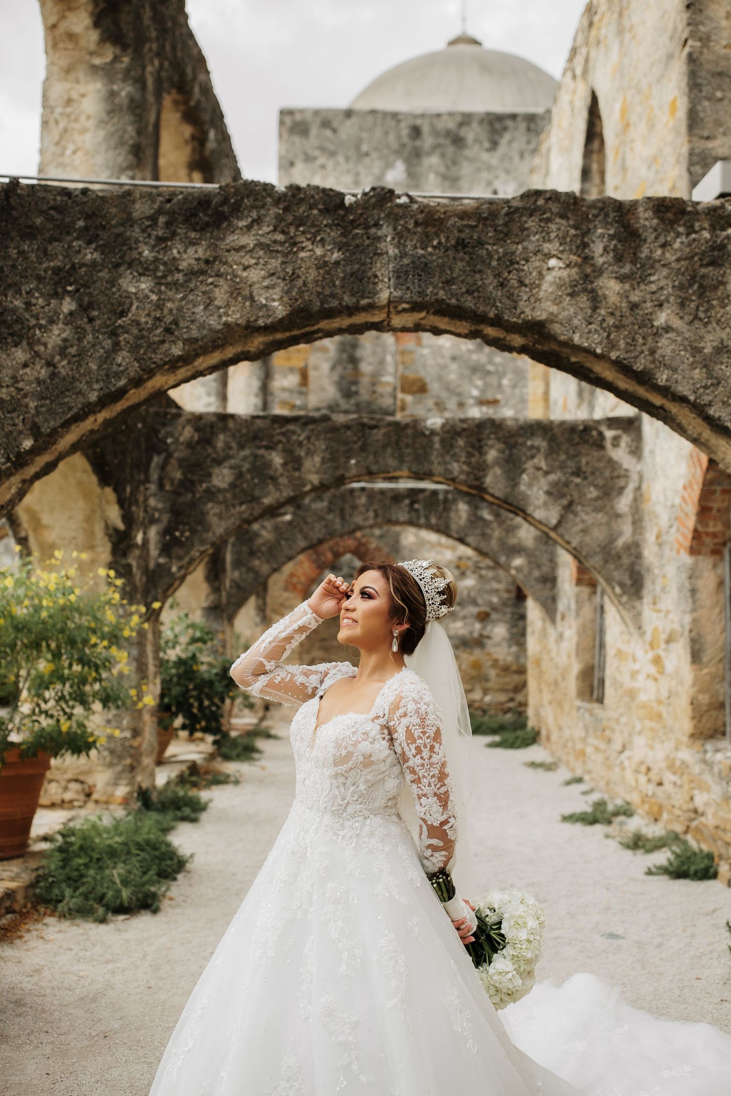 Bride in lace ball gown with crown poses at historic Mission San Jose stone archways in San Antonio.