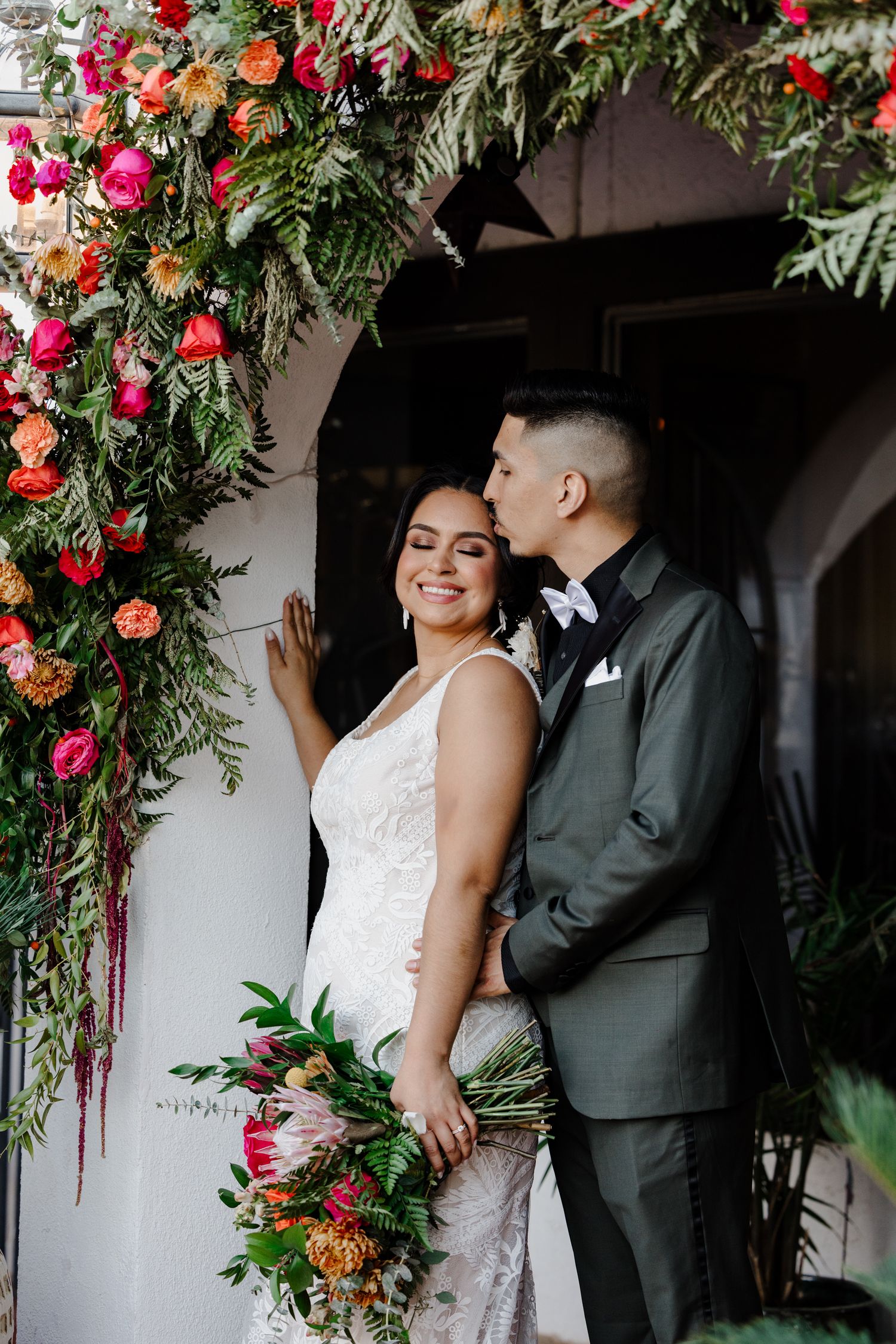 Wedding couple poses under beautiful floral arch with red and orange blooms.