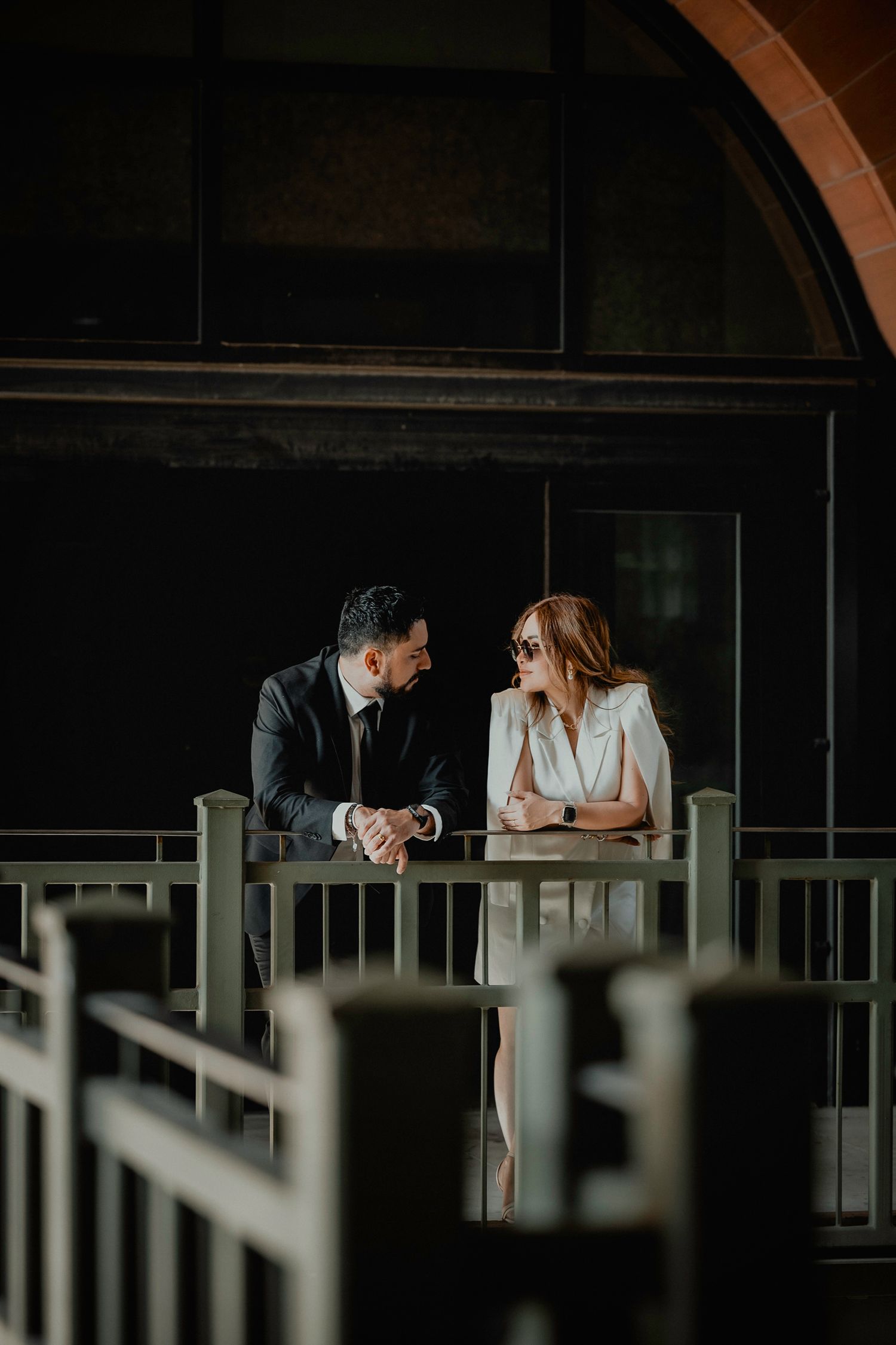 A stylish couple in formal attire shares a conversation on a dark wooden balcony.
