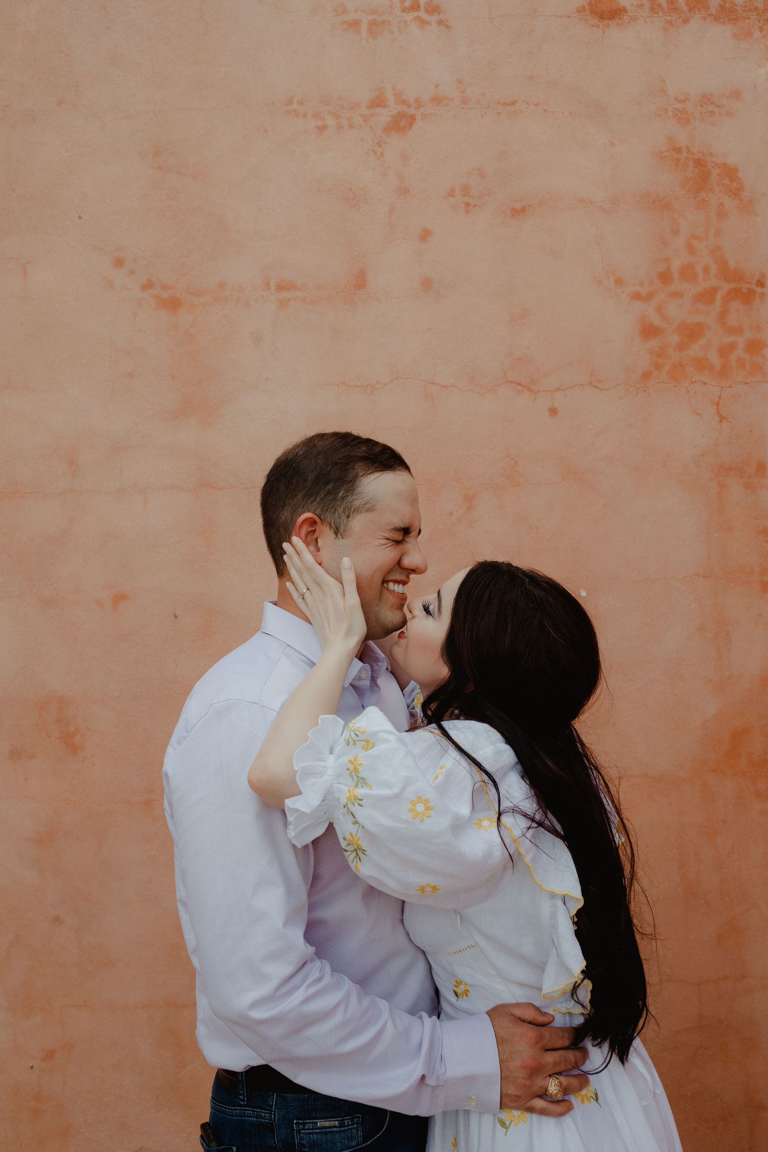 Smiling couple about to kiss against a warm terracotta wall outdoors.