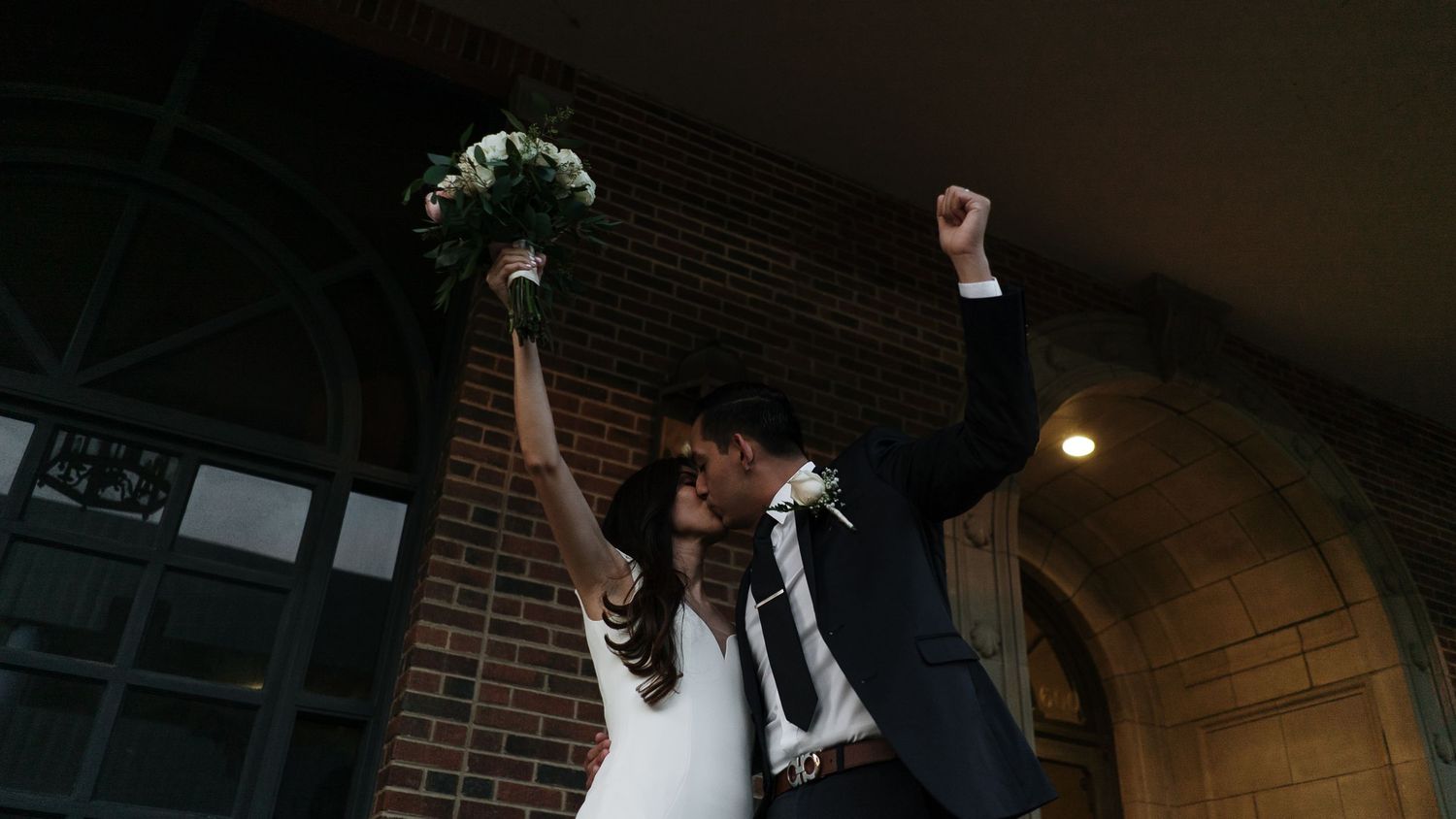 Newlyweds celebrating with raised arms and bouquet outside brick wedding venue.