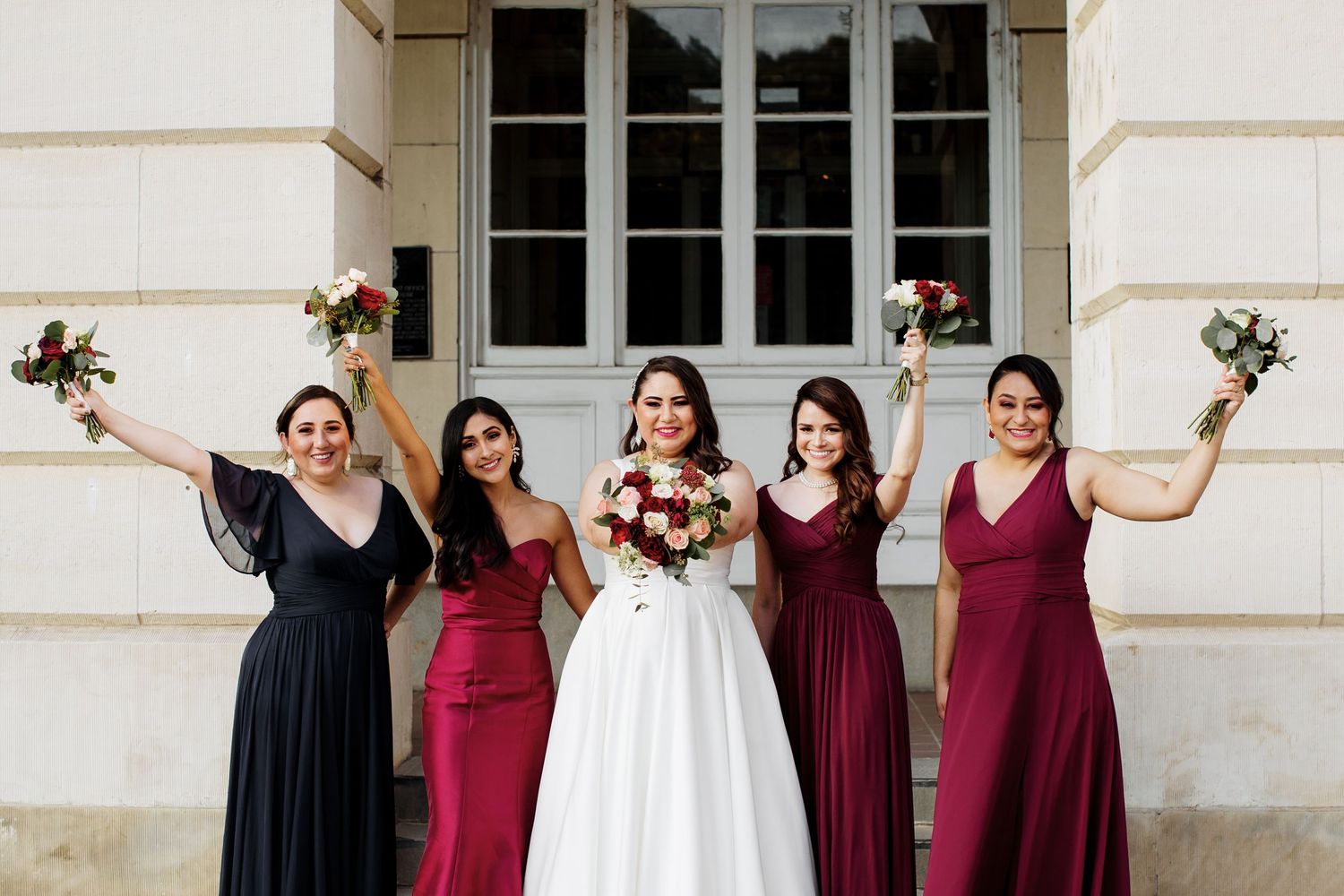 Bride posing with four bridesmaids in burgundy and navy gowns holding bouquets outside a building.