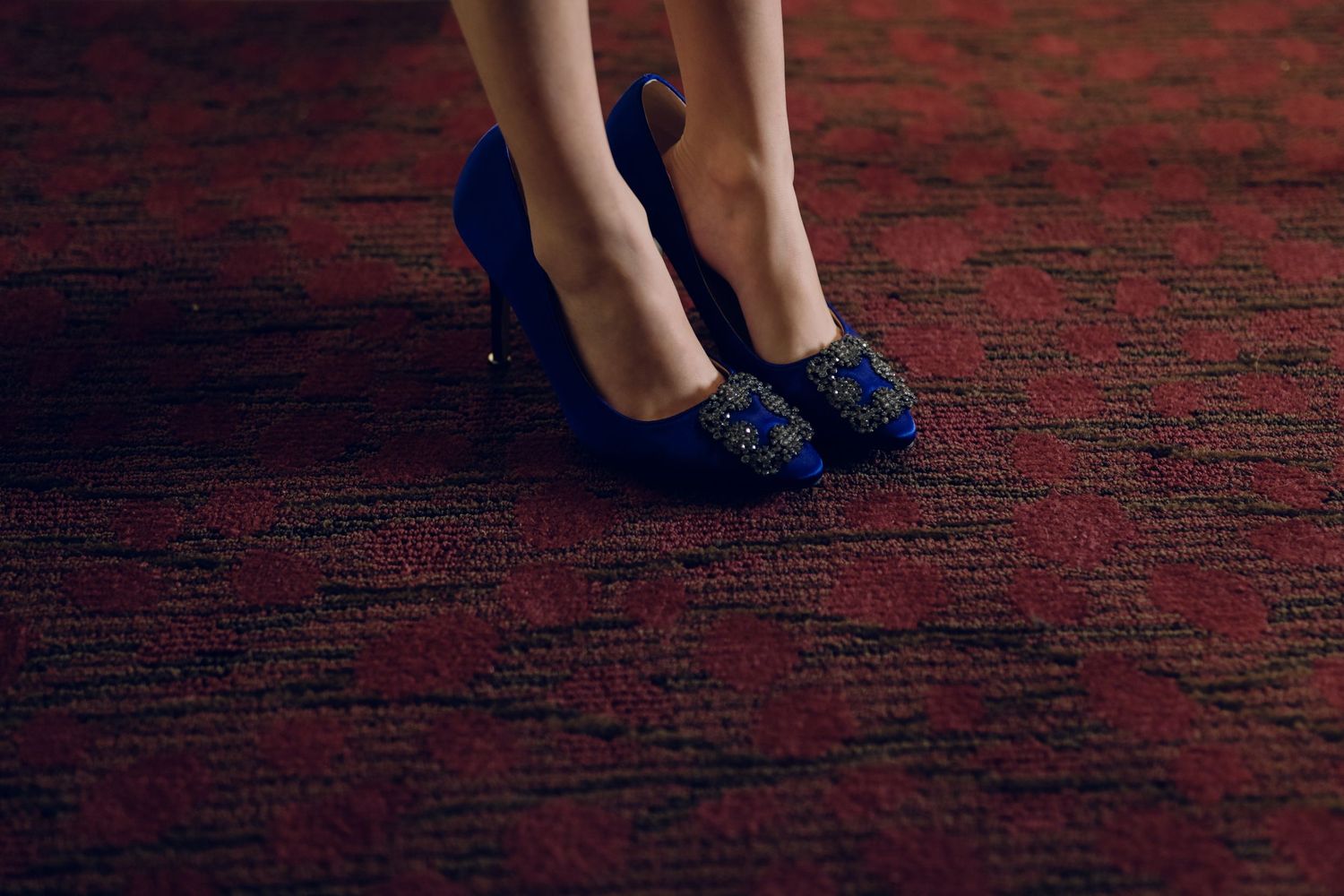 Bride wearing ornate royal blue embellished heels standing on red patterned carpet at wedding.