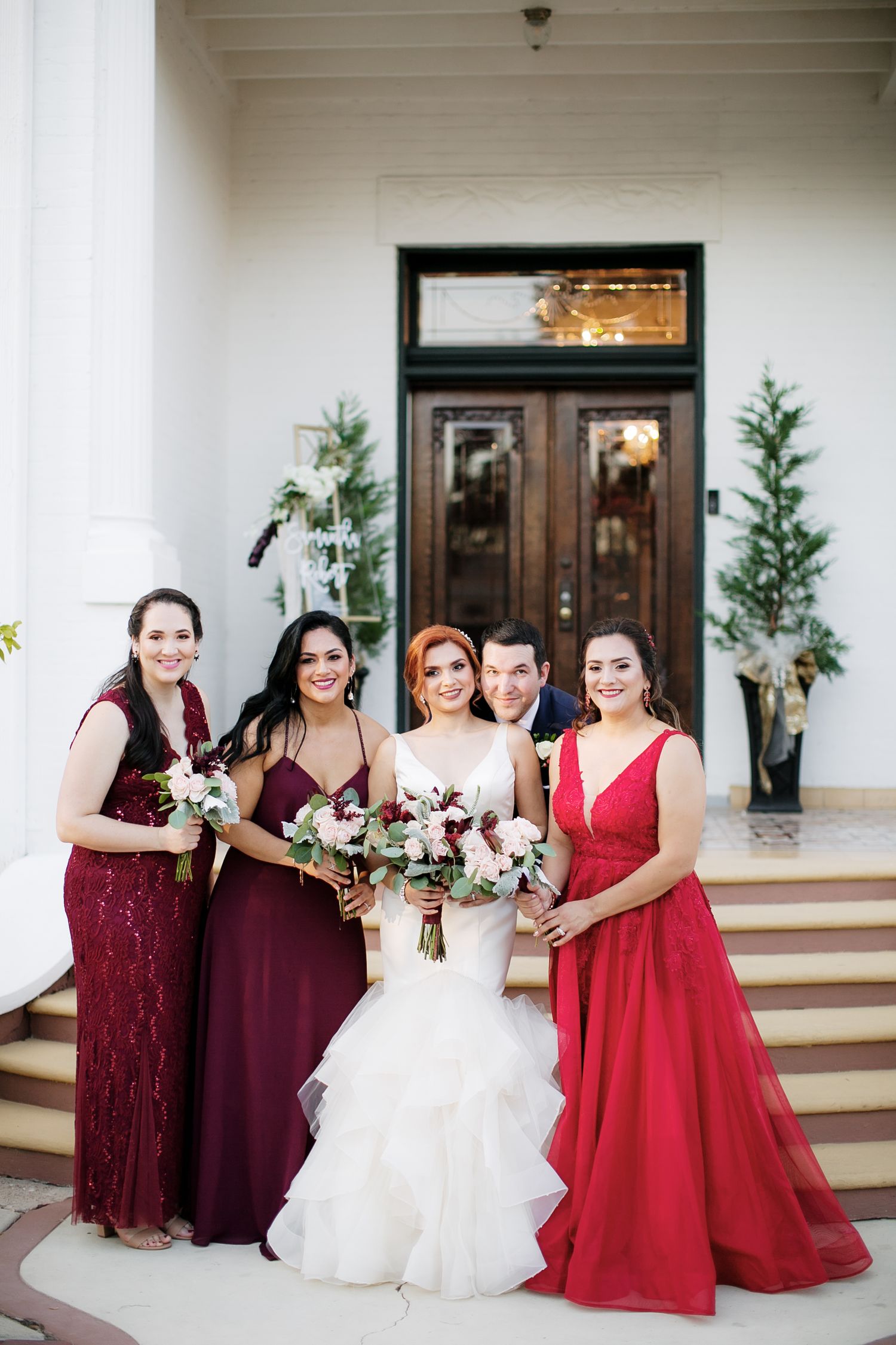 Bride in white gown poses with bridesmaids in burgundy and red dresses holding bouquets outside elegant venue doors.