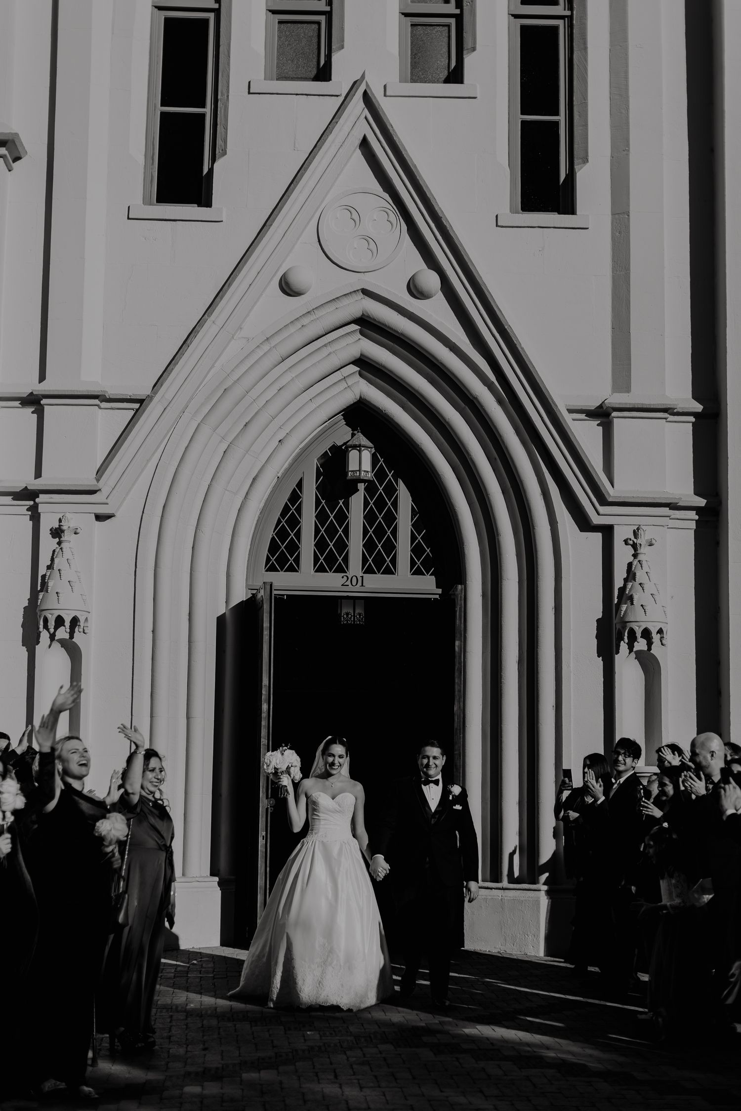 Bride and groom exit a Gothic church doorway in black and white wedding photo with guests celebrating outside.