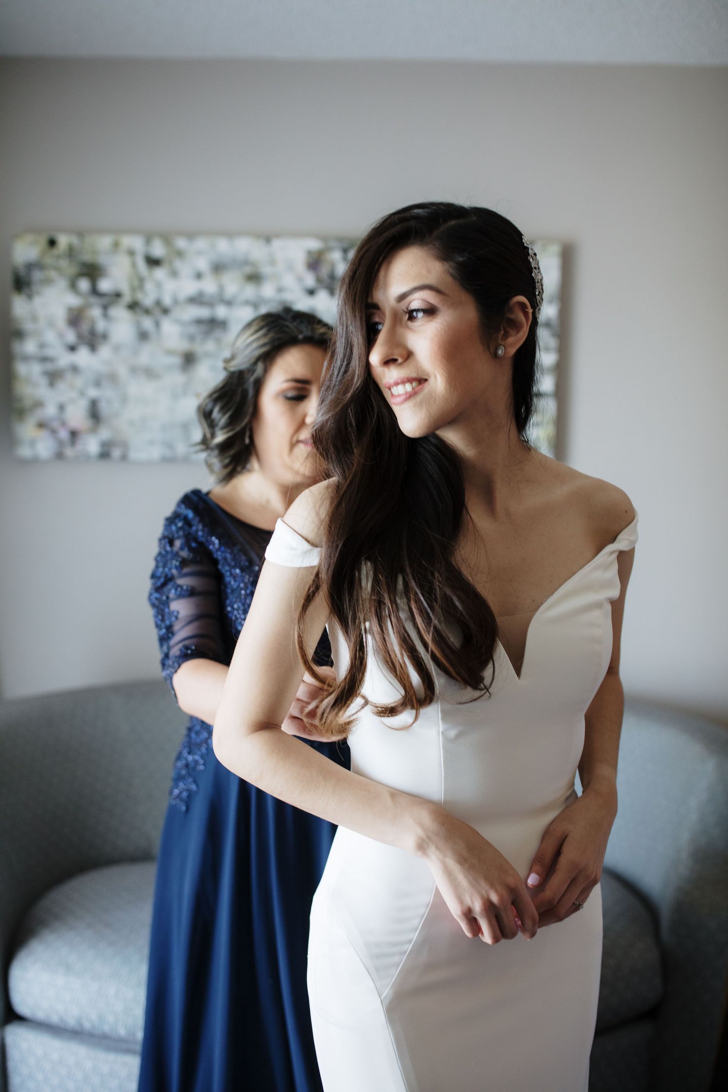 A smiling bride in a white off-shoulder wedding gown gets help dressing from a woman in a blue lace dress.